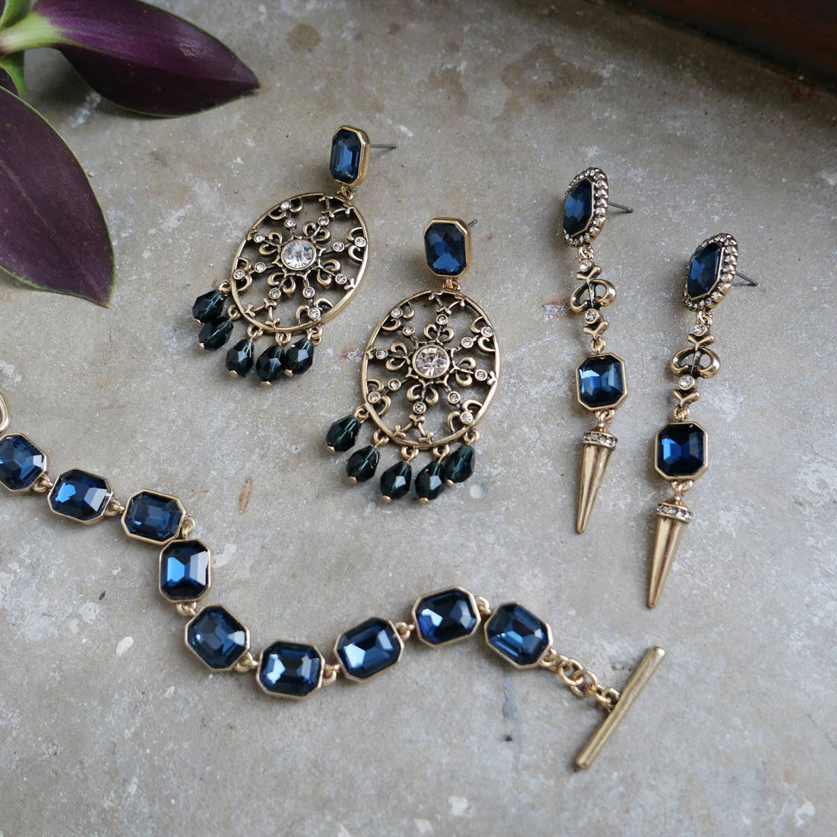 The A&C Oslo Antoinette Midnight Blue Statement Earrings, gold plated and featuring a bold design, are displayed on a stone surface beside purple leaves.