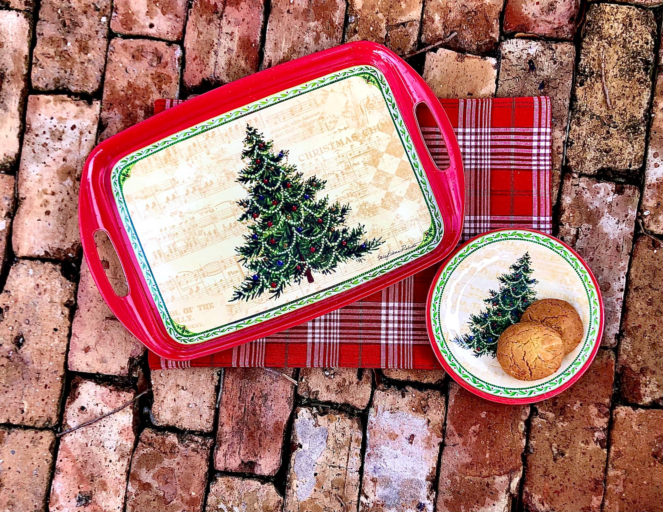 The Merry Christmas Tree Bamboo Serving Tray rests on a plaid cloth; the tray, adorned with festive tree designs and holding cookies, creates a charming hostess gift for Christmas on a brick surface.