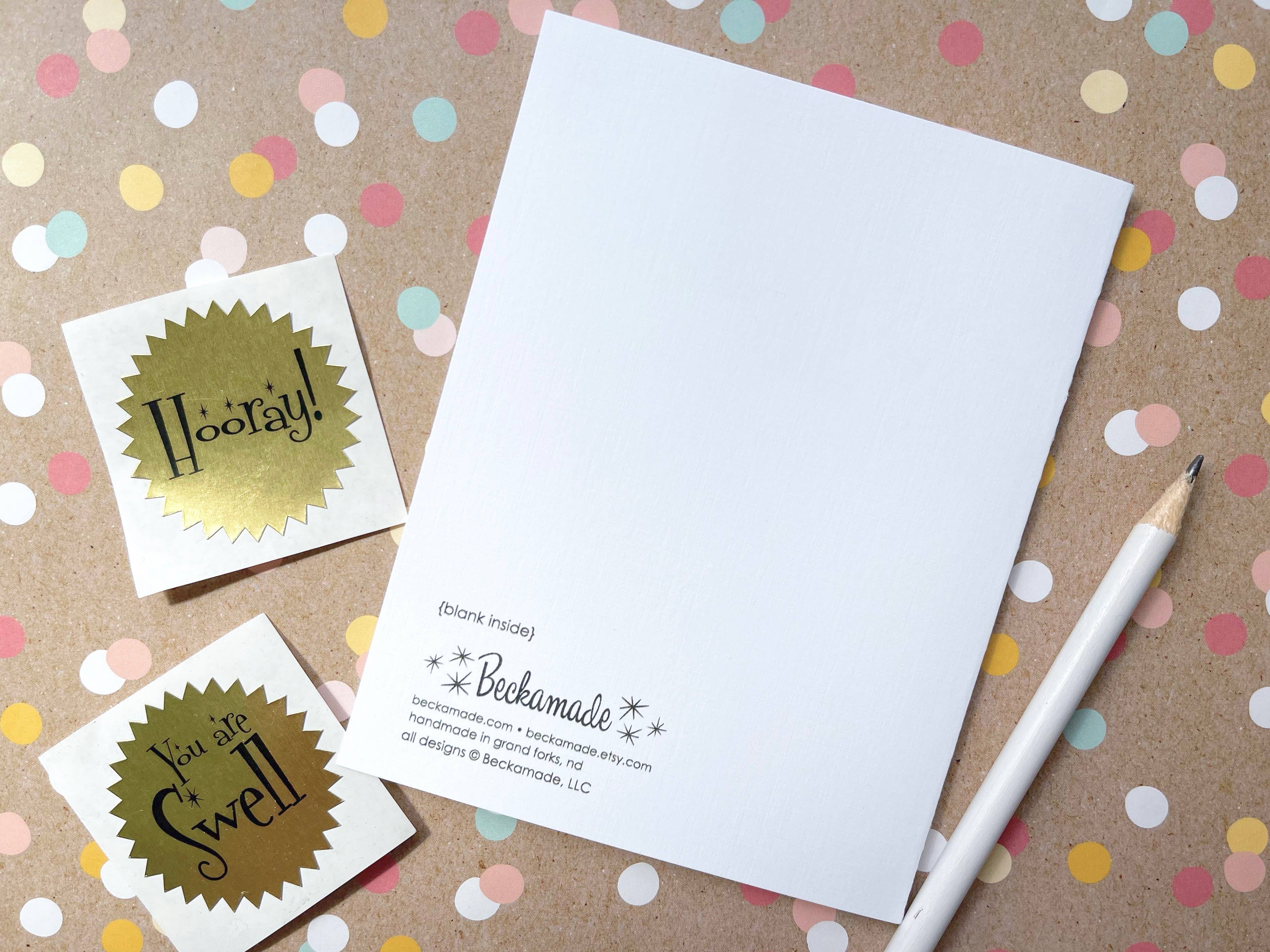 Uffda Floral Card on a confetti-patterned background features two gold stickers with the messages “Hooray!” and “You are Swell.” Printed on elegant linen texture card stock, this card comes in a biodegradable sleeve. A white pencil is positioned to its right. The interior of the card is blank, awaiting your personal touch.