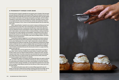 A hand sifts powdered sugar over three cream-filled Danish pastries on a dark surface. To the left lies "Book: Scandinavian from Scratch" by Penguin Random House, open to a page about the pastries' history in Scandinavian baking.