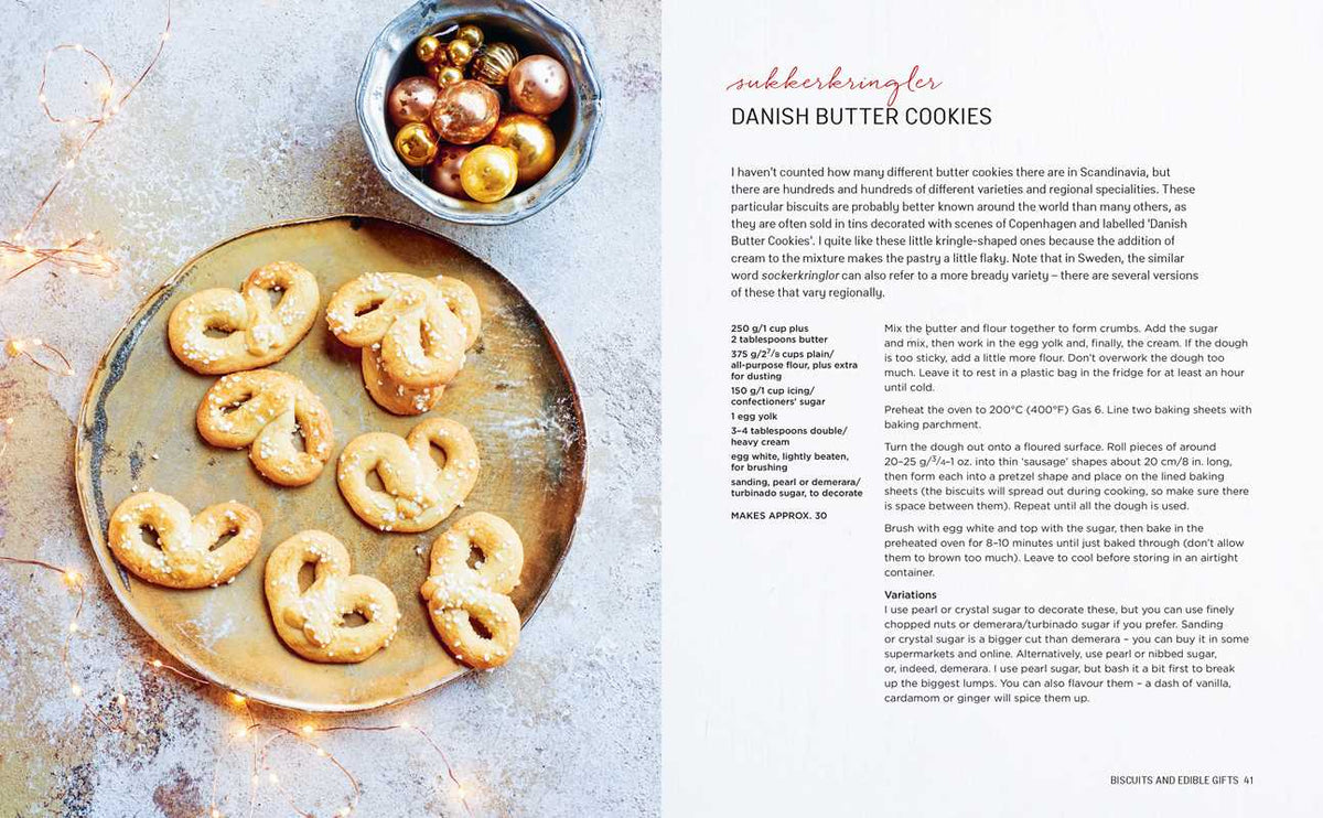 Seven Danish butter cookies with pearl sugar on a gold tray, next to festive ornaments on a white surface.