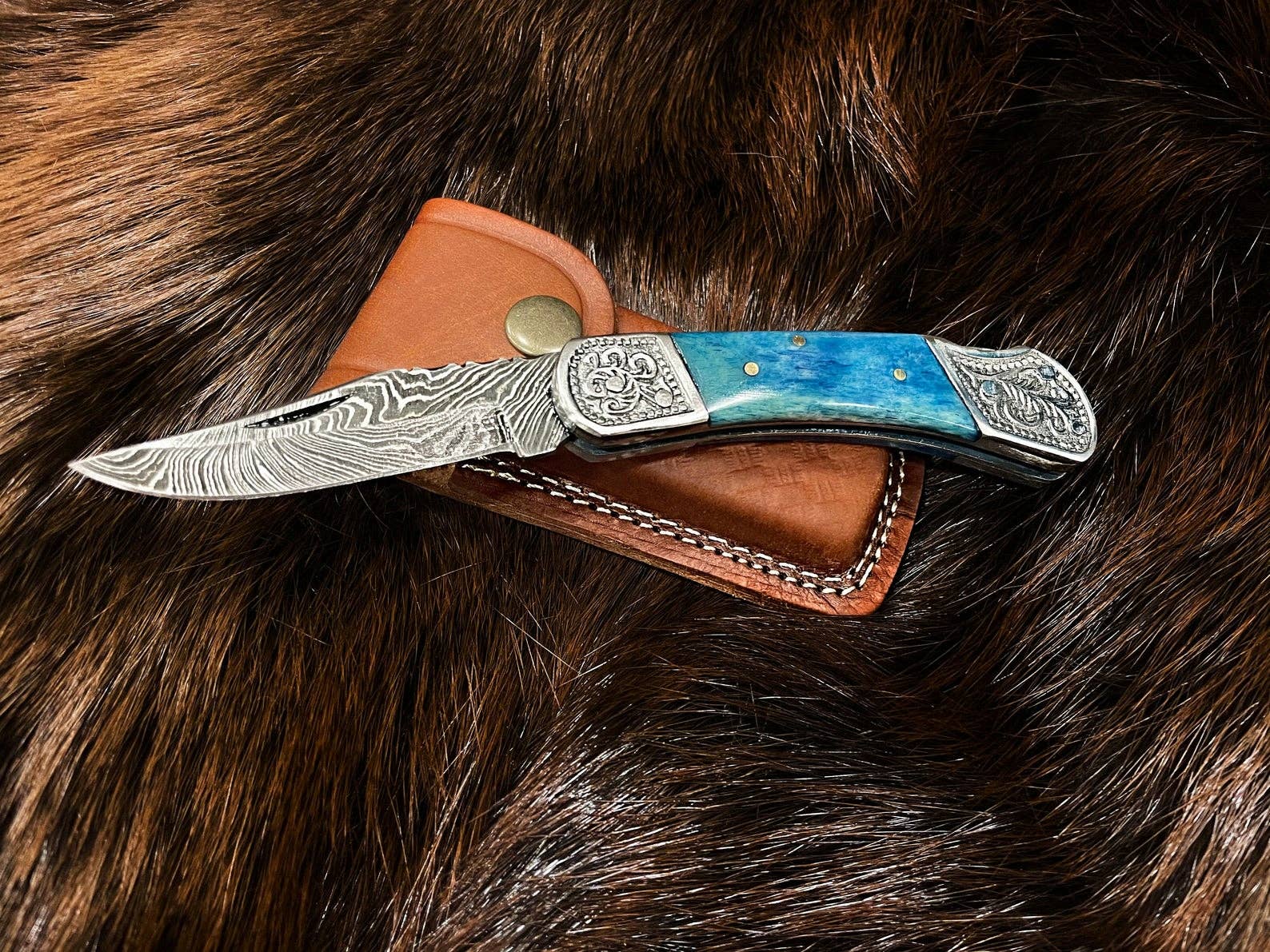 The Knife: Dyed Blue Bone 6.5 Damascus Steel features an engraved Damascus blade and a blue handle, displayed on a brown leather sheath atop fur.
