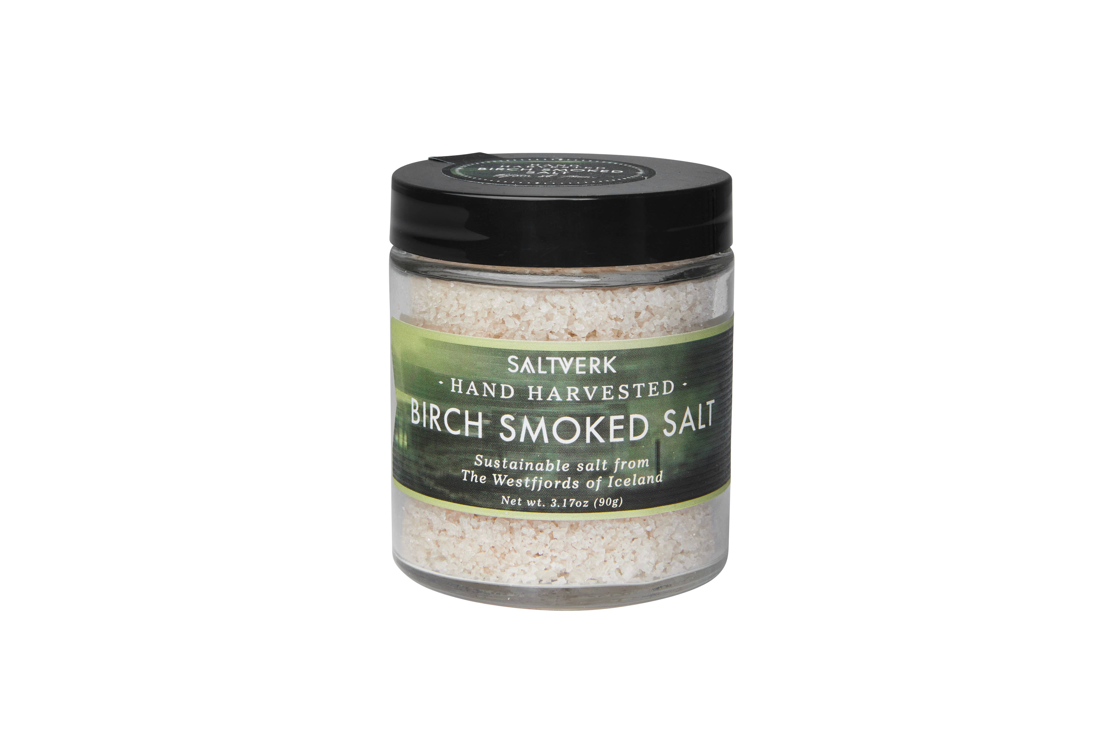 A clear jar from the SALTVERK Gift Set features hand-harvested Icelandic Birch Smoked Sea Salt, with a green label and black lid. The set includes Flaky Sea Salt, Lava Salt, Birch Smoked Salt, and Arctic Thyme Salt from the Westfjords.