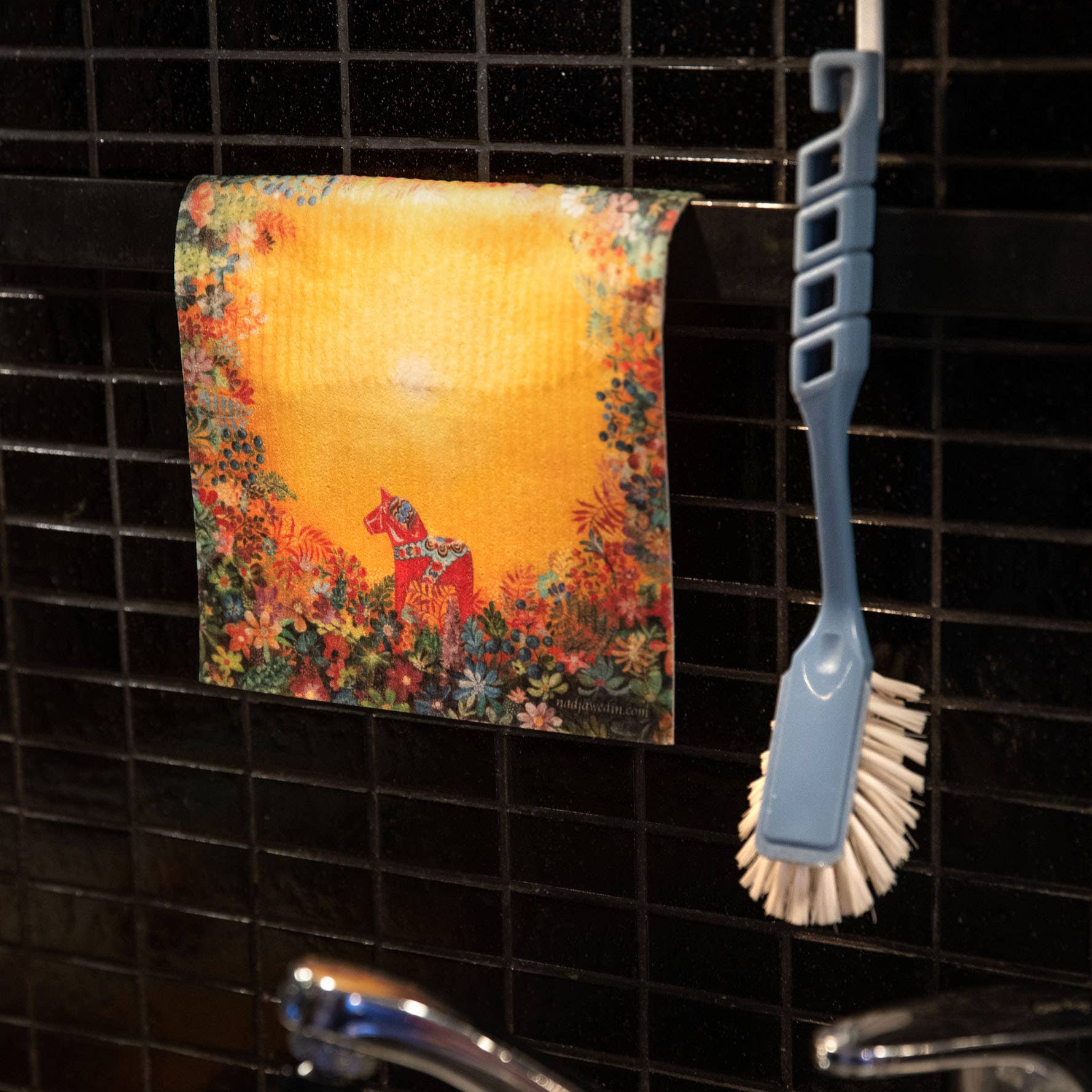 The Dish Cloth: Light of Grytnäs Dala Horse, featuring a vibrant floral and horse design, hangs on a black tiled wall beside a blue dish brush—an eco-friendly, charming alternative to traditional towels.