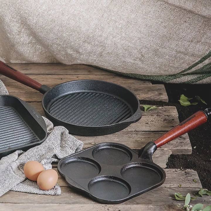 Three Skeppshult 8 egg pans with walnut handles rest on a rustic wooden surface, joined by two brown eggs and a textured cloth in the background.