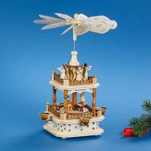 The Christmas Pyramid: Nativity Square 13" Tall features two tiers with nativity figurines and angels, topped with a spinning fan. Elegantly crafted, it stands against a blue background with a fir branch and glowing candles.