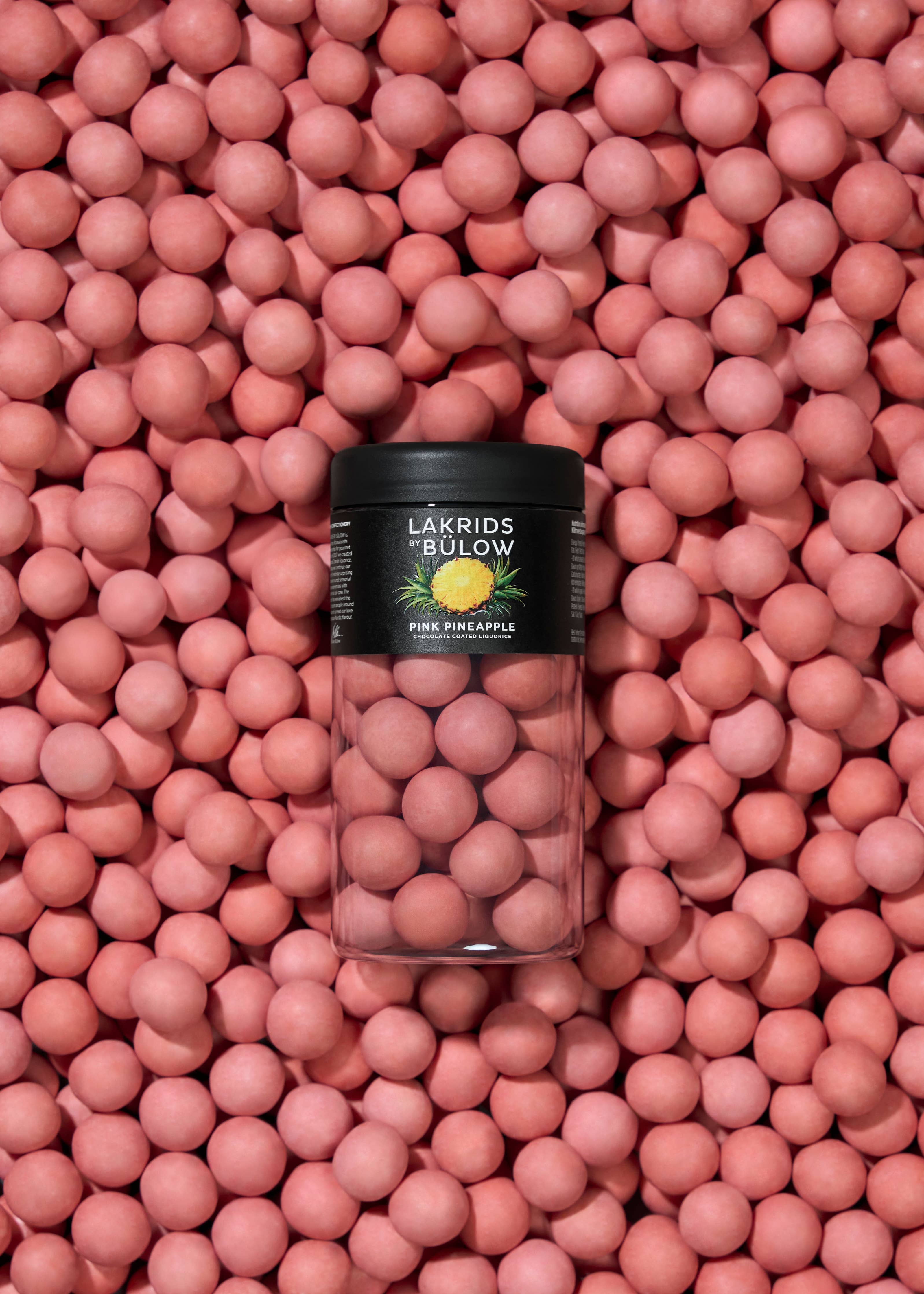 A clear jar of Lakrids by Bülow - Pink Pineapple Danish Chocolate Coated Licorice (125g) sits on a surface covered with the same pink licorice candies.