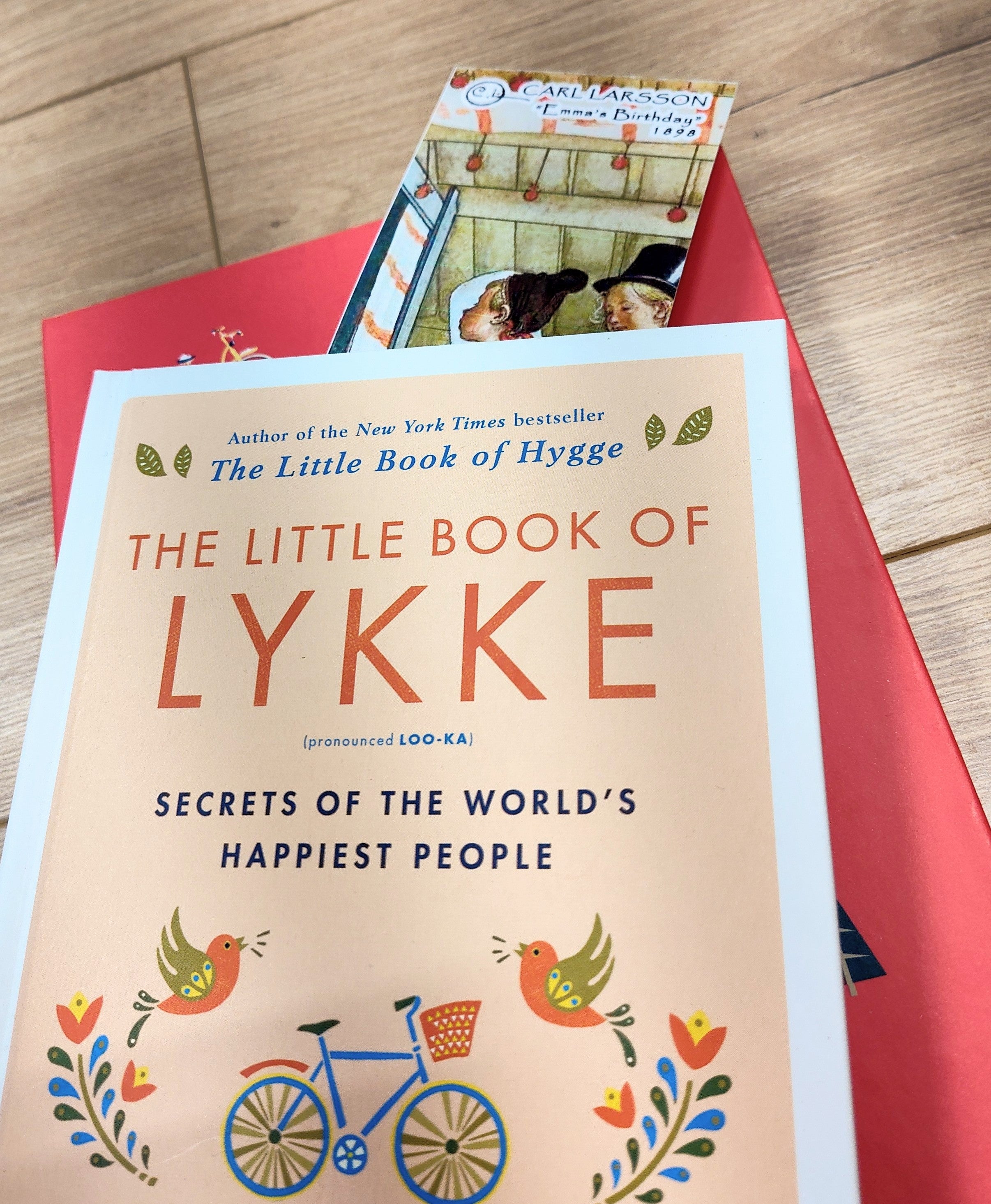 A copy of The Little Book of Lykke rests on a red folder, with the Bookmark: Carl Larsson “Emma’s Birthday” (8.5" × 2.5") partially visible between its pages on a wooden surface.