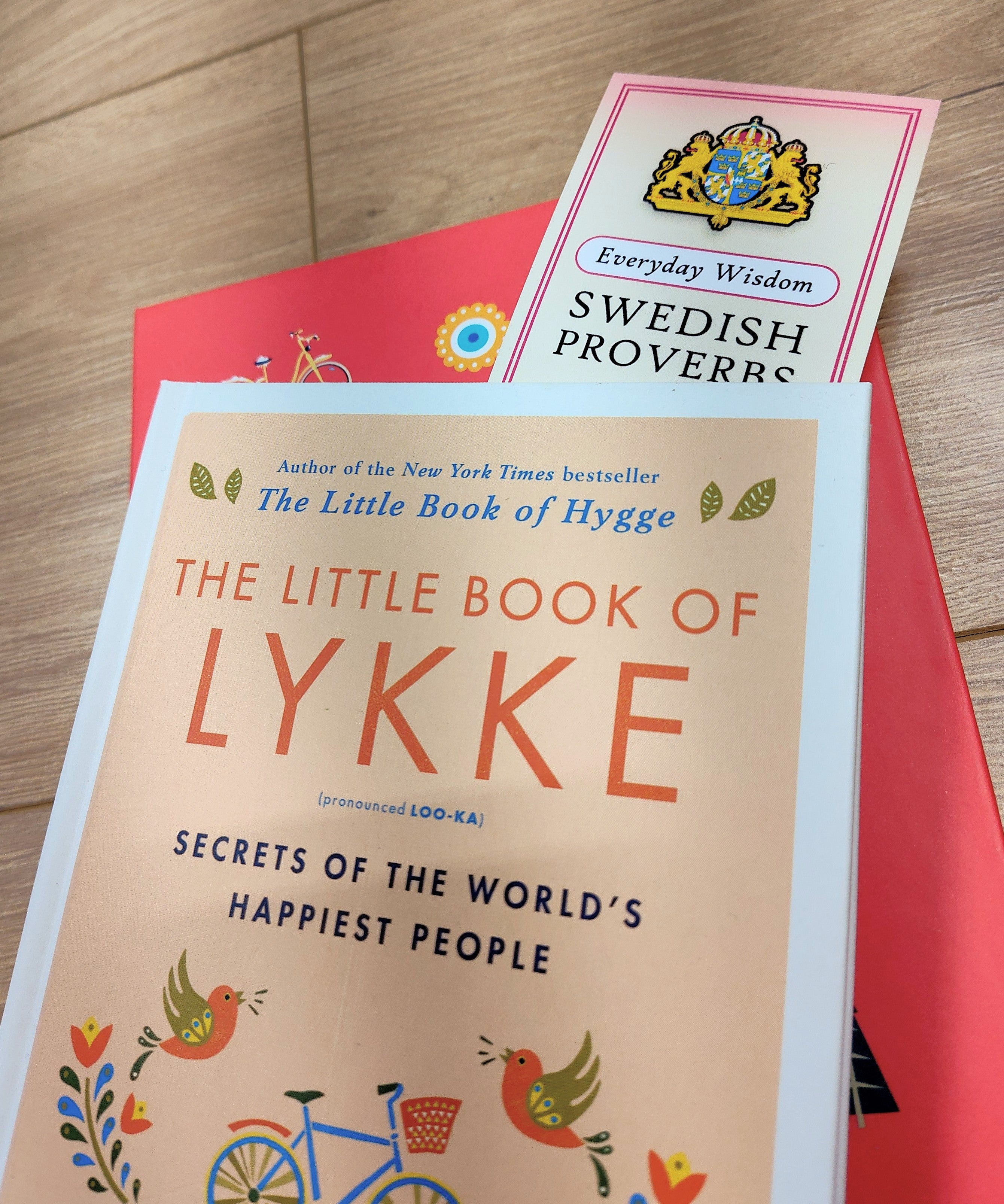 A close-up of The Little Book of Lykke on a red notebook, with the Bookmark: Swedish Proverbs “Everyday Wisdom” (2.5" × 8.5") partially visible—all atop a wooden surface, evoking Scandinavian wisdom.