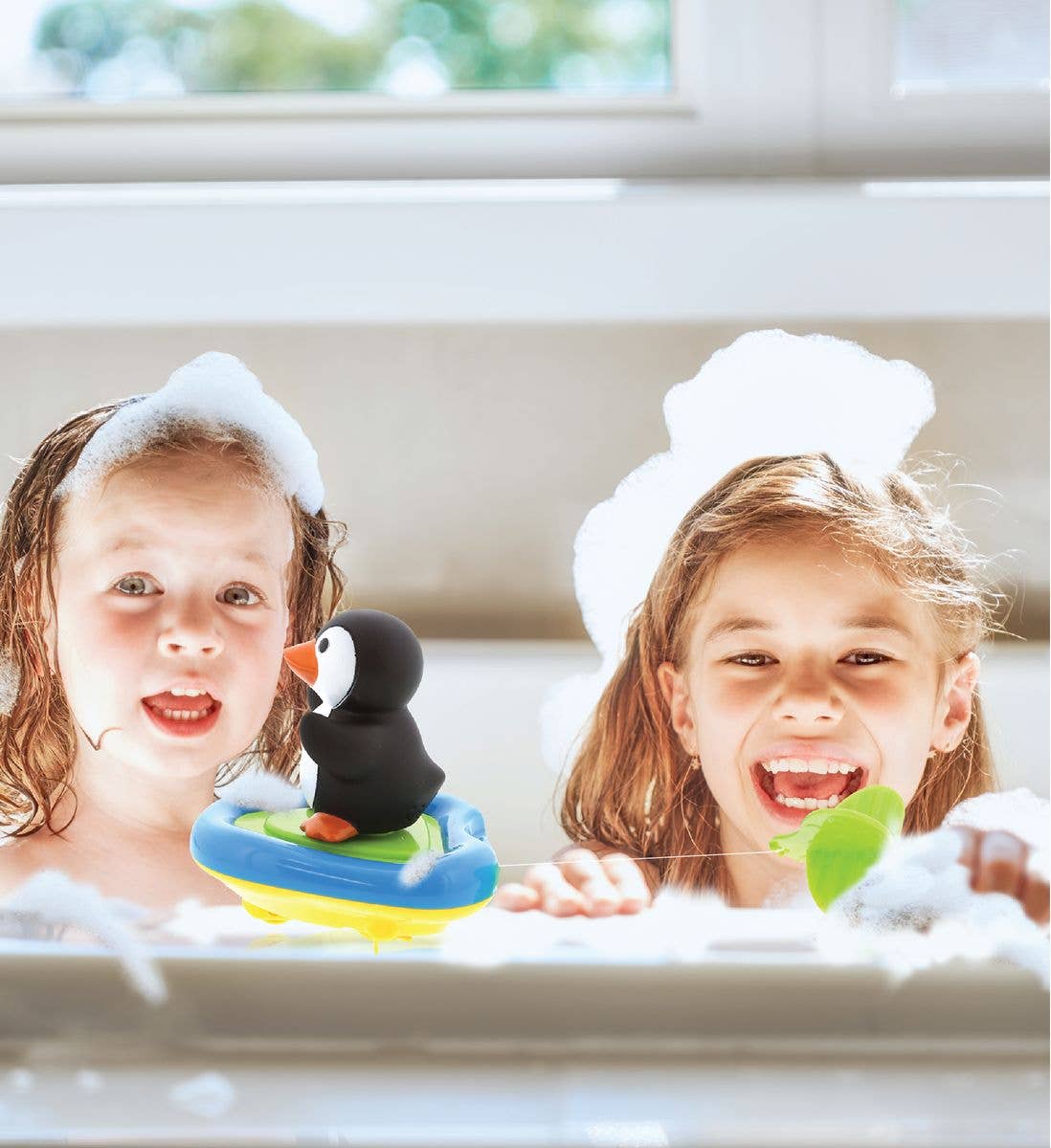 Two children in a bubble bath smile at the camera, foam on their heads, while the Bath Toy: Penguin Boat Racers Buddy floats nearby.