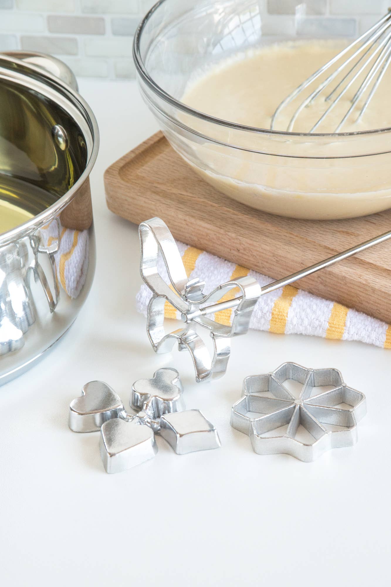 The Cookie Cutter: Rosette Set, Cast-Aluminum, 4-Piece Set sits beside a saucepan and bowl of batter with a whisk—perfect for making Scandinavian rosette cookies. A wooden cutting board and striped towel add to the cozy baking scene.