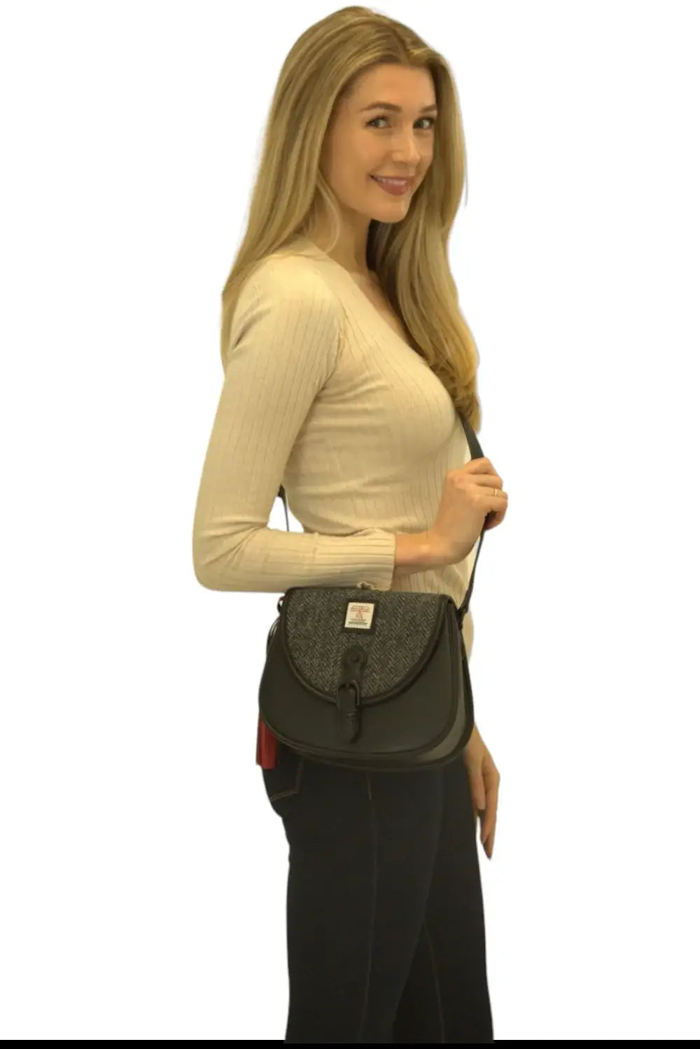 A woman with long blonde hair, wearing a beige ribbed top and black pants, poses sideways holding the Harris Tweed Saddle Bag - Grey against a plain white background.