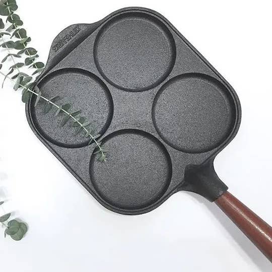 The Skeppshult Egg Pan With Walnut Handle 8 features four circular molds in a square cast iron design, shown on a white surface next to a eucalyptus branch.