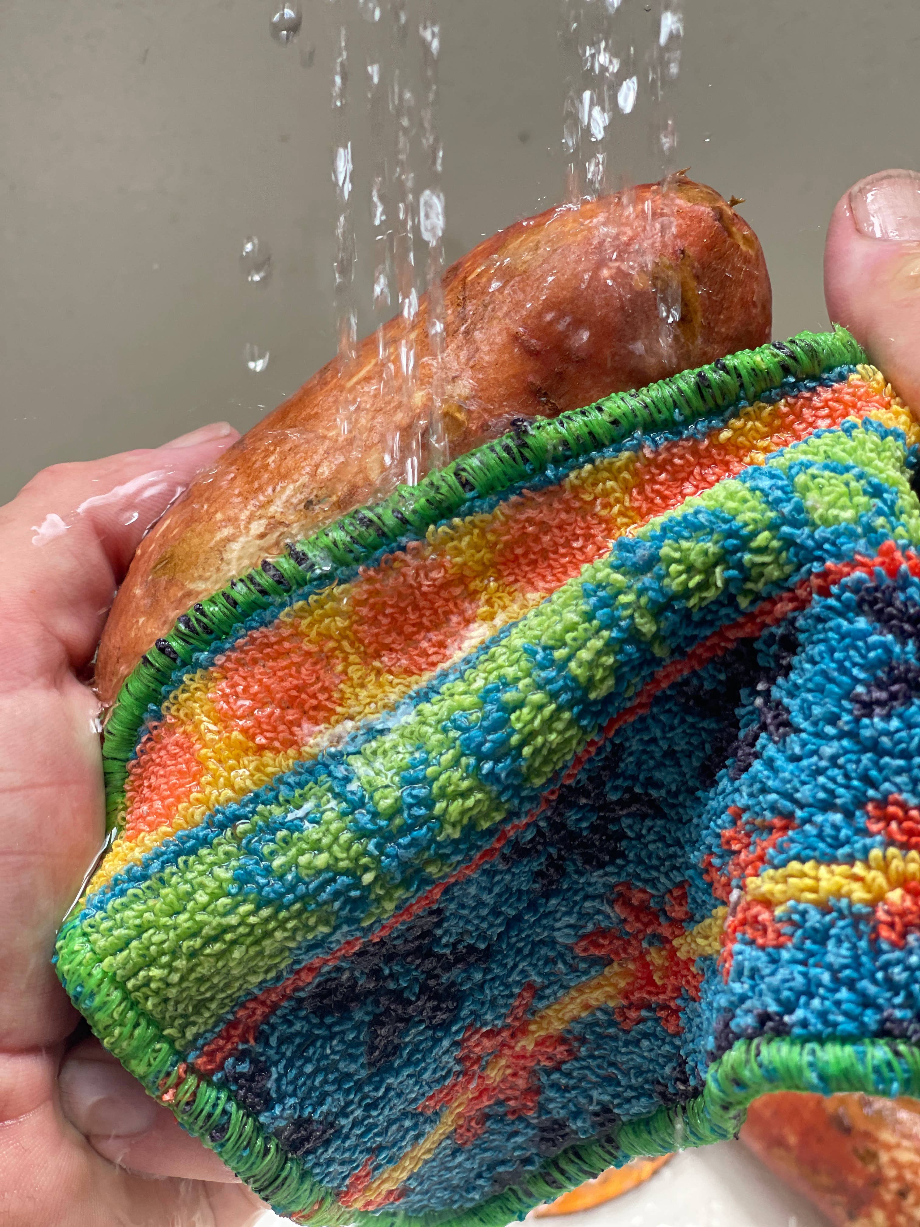 Someone uses the Scrubby: Vogue Skrubba Cloth, a colorful, scratch-free scrubber, to wash a sweet potato under running water.