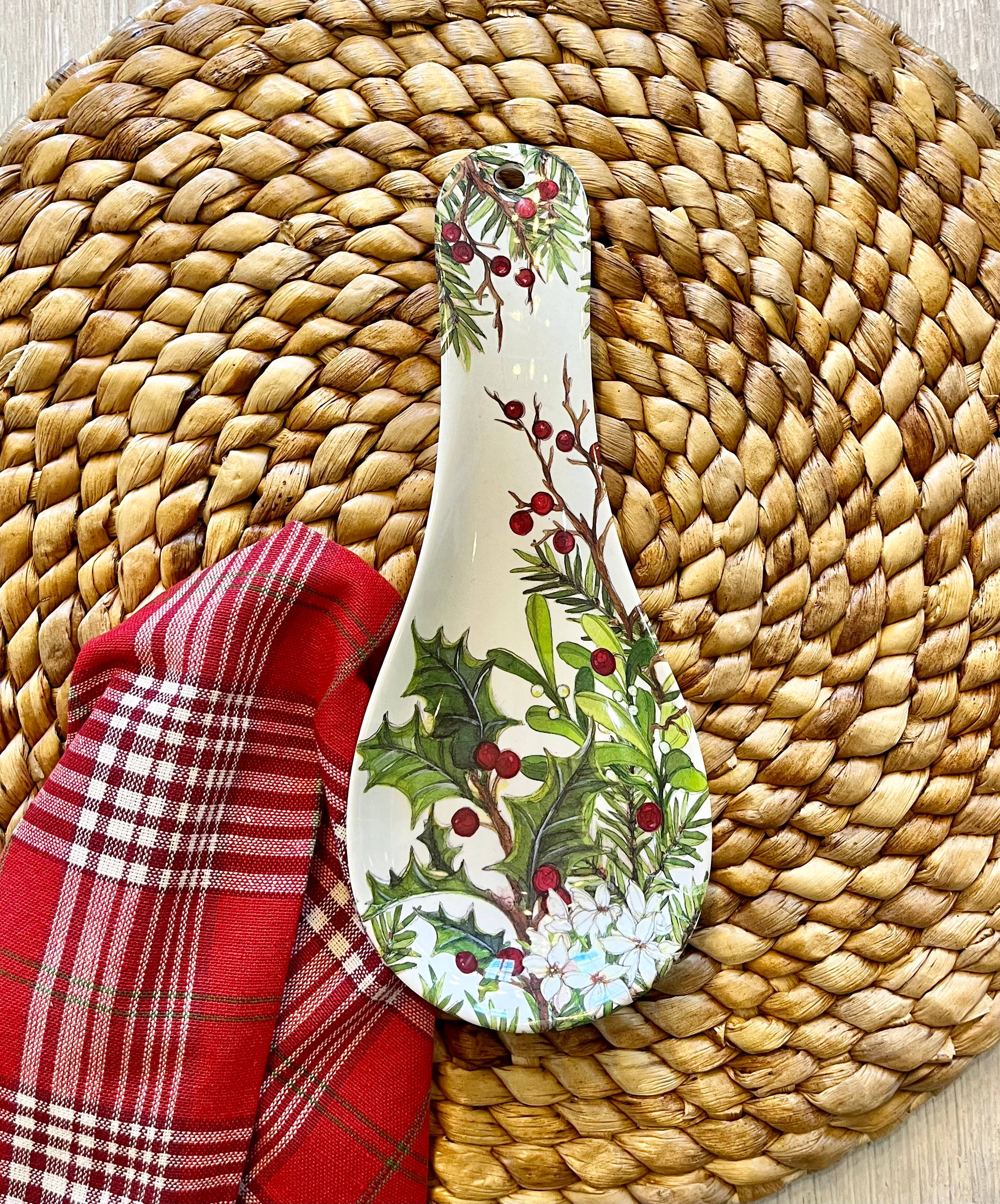 The Spoon Rest: Holly & Berries Christmas Holiday features a festive design and sits on a woven placemat next to a red plaid cloth napkin.
