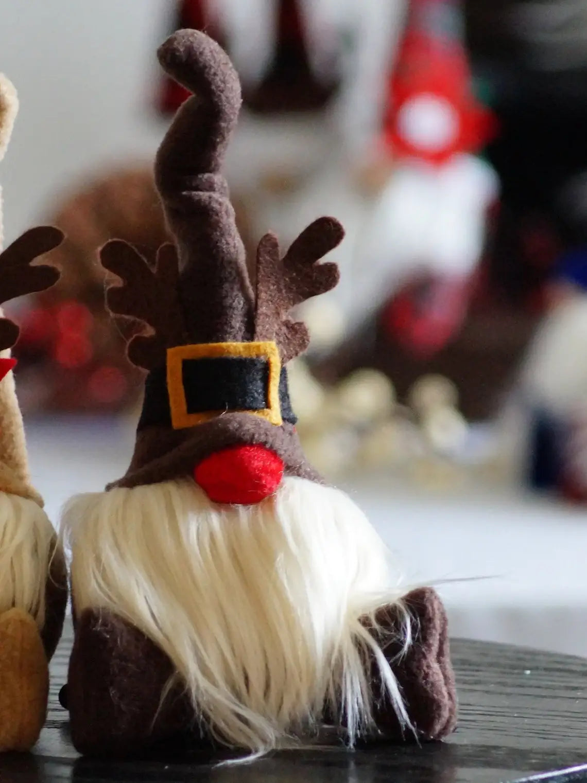 The Gnome: Rudolph Gnome 9" Tall Sitting features a long, twisted brown hat, faux reindeer antlers, a red nose, and a white beard—perfect for adding festive spirit to your Christmas decor.