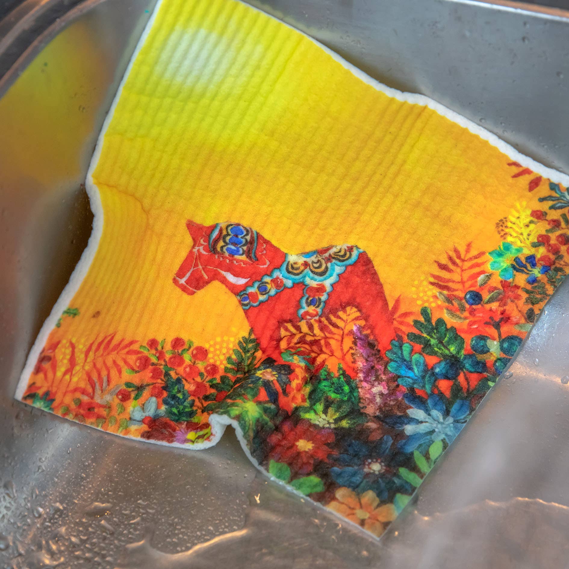 The Light of Grytnäs Dala Horse Dish Cloth, adorned with a red horse and vibrant floral design, rests effortlessly in the stainless steel sink.