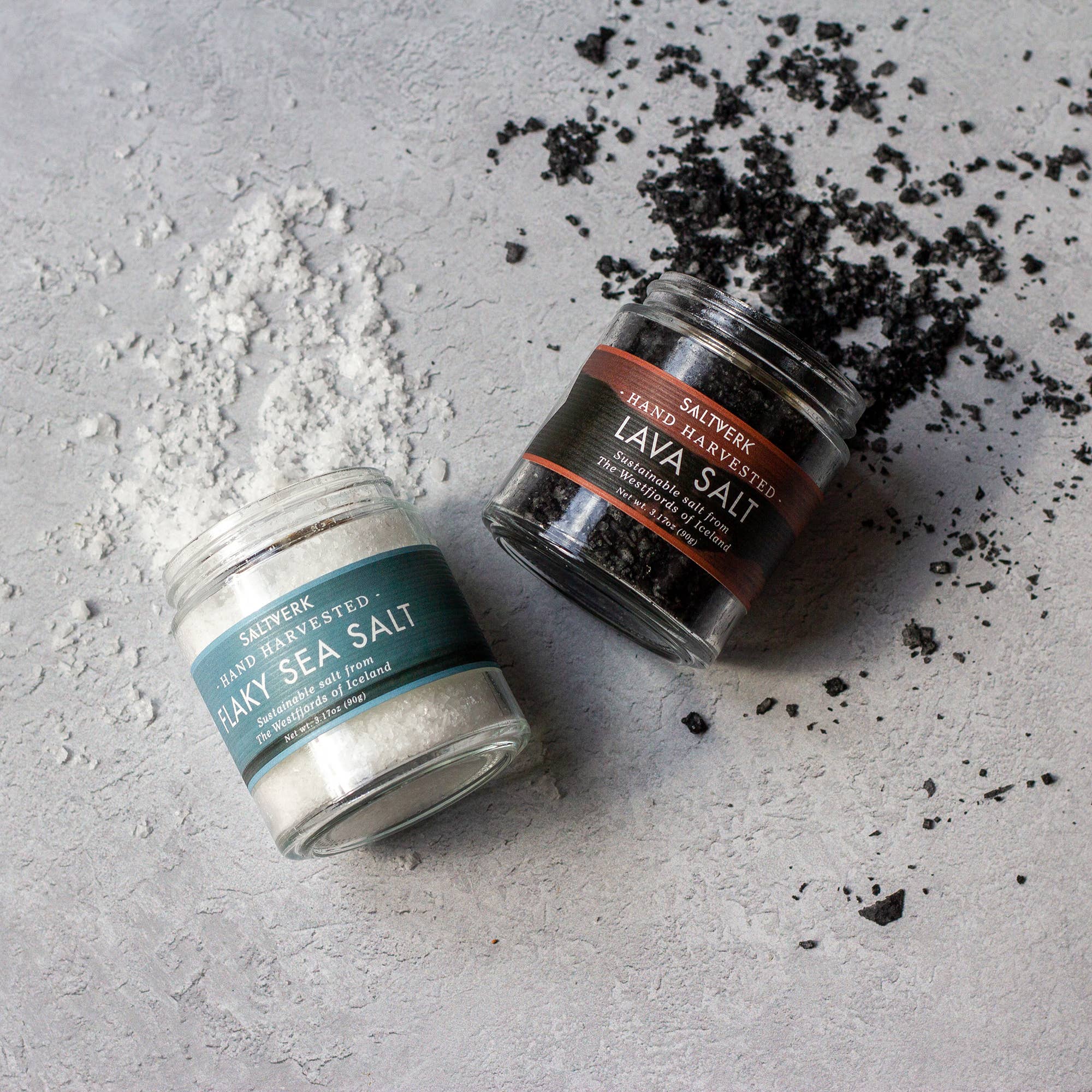 The SALTVERK Gift Set features two glass jars, one with Icelandic sea salt and the other with black lava salt, tipped on a gray surface with spilled salt around them.