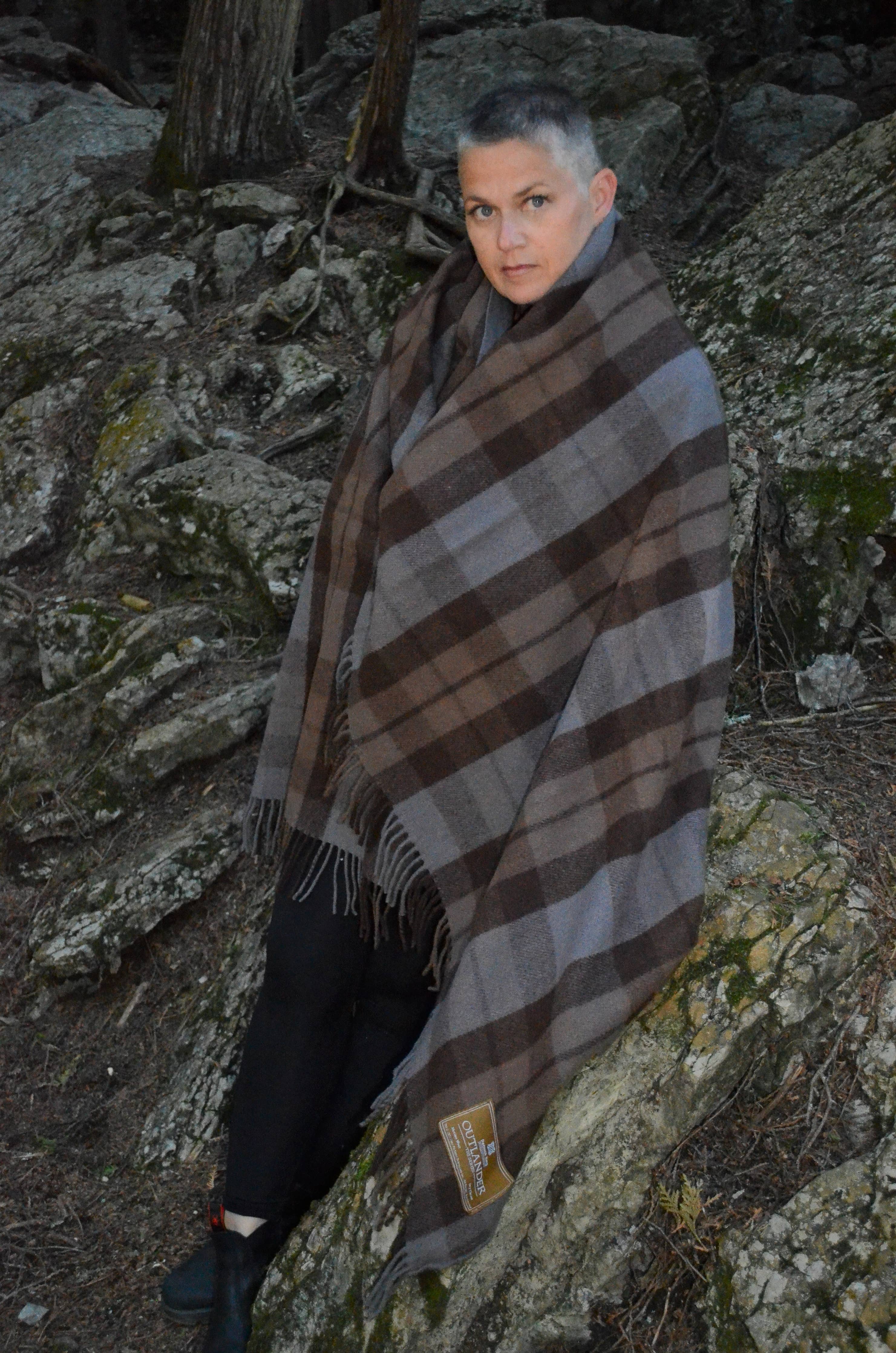 A person with short hair stands outdoors on rocky terrain, wrapped in a large brown and gray Outlander Merino Wool Blanket, looking at the camera.