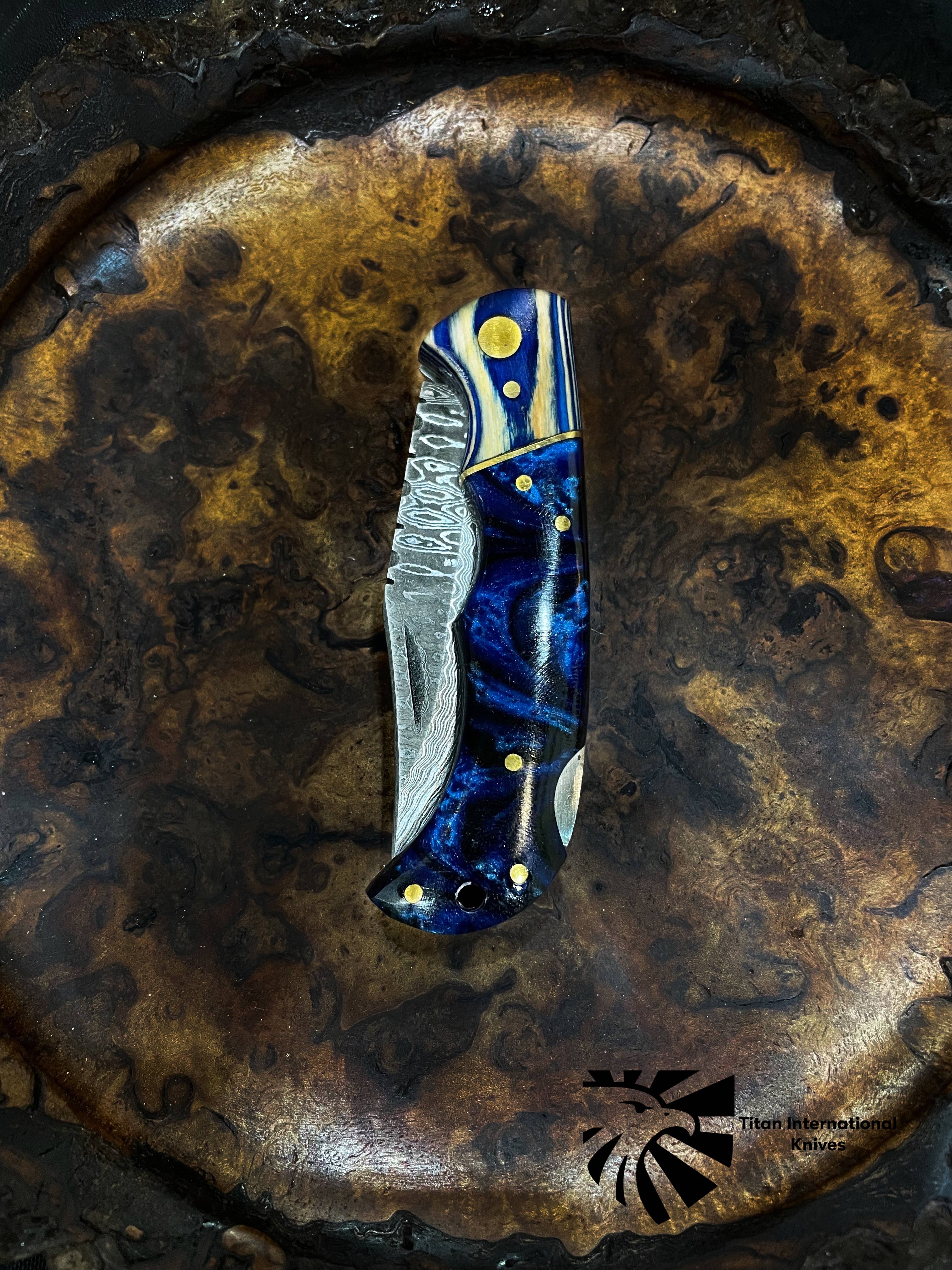 The Titan Celestial Damascus Pocket Knife, featuring a blue and gold handle and patterned hand-forged blade, rests on a textured brown surface. The striking detail includes the Titan International Knives logo in the lower right corner.
