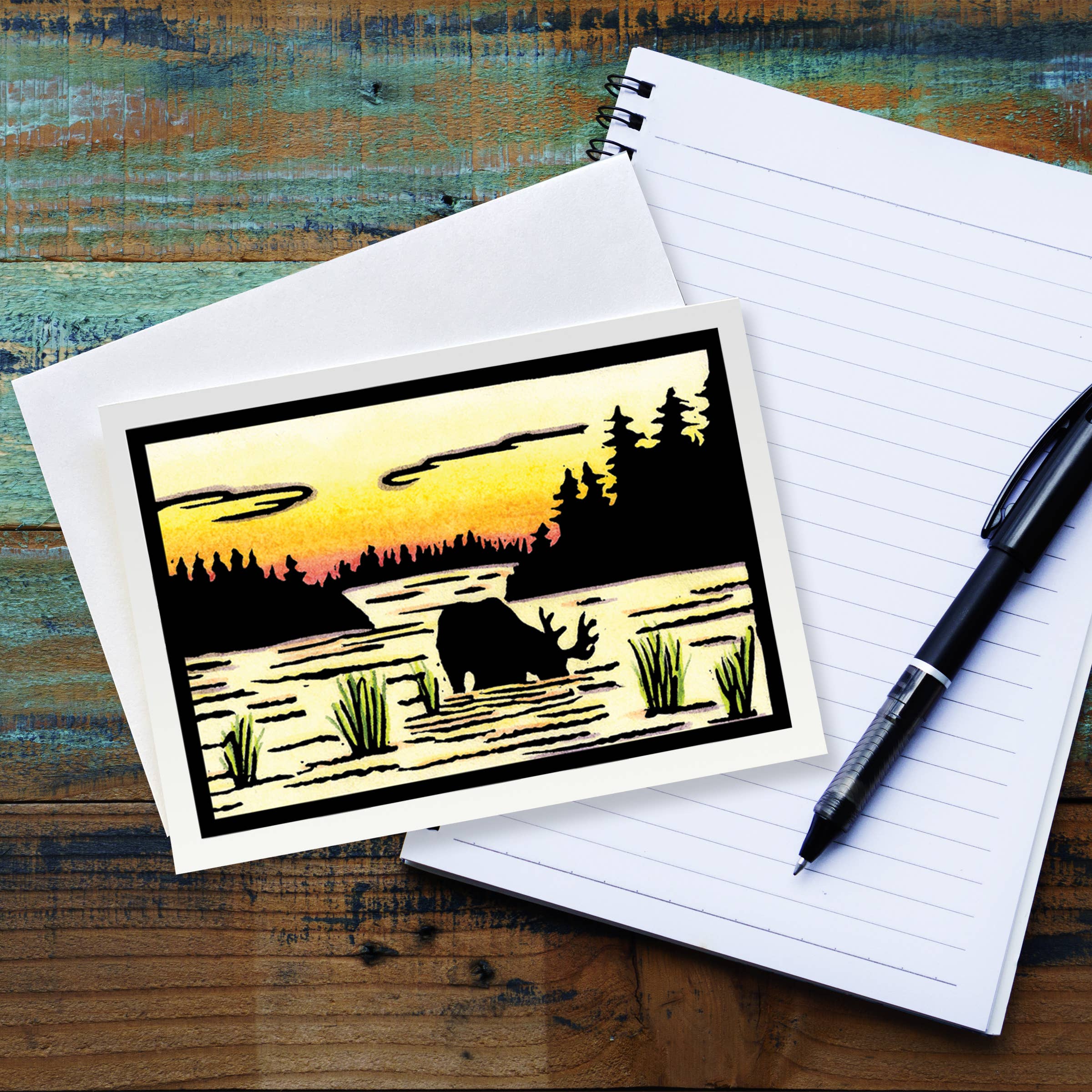 The Card: Evening Drink Moose by Sarah Angst, a 5x7 greeting card with envelope (blank inside), features a moose and forest silhouette at sunset and rests on a lined notepad with black pen atop a rustic wooden surface.