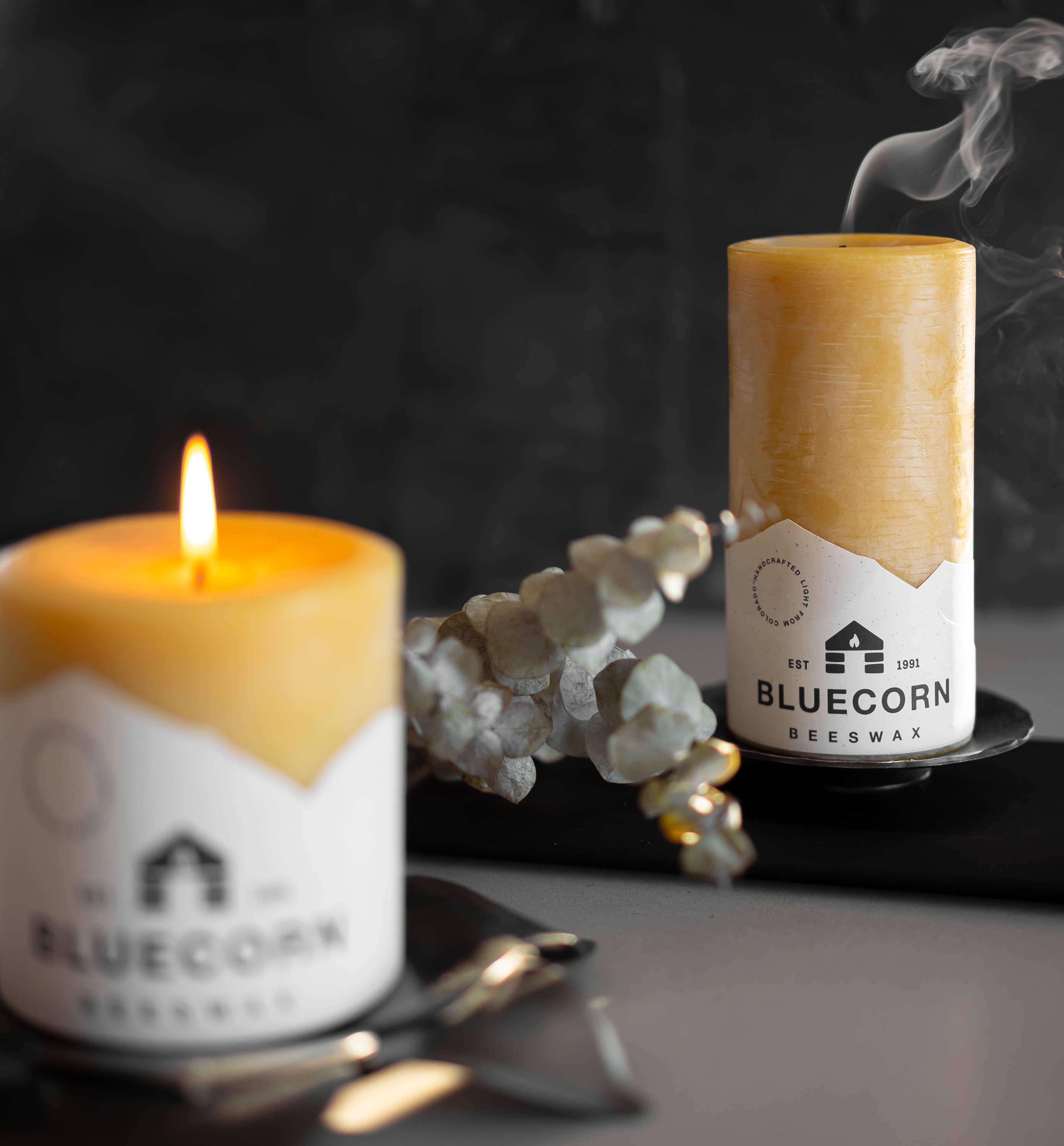 Two Bluecorn beeswax candles rest on a table in a Forged Steel Pillar Candle Base (4.75" diameter); one glows brightly while the other, just blown out, releases a gentle wisp of smoke.