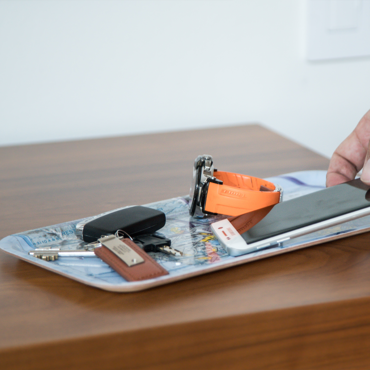 Birchwood tray on a wooden surface with keys, orange-strap watch, phone, pen, and black case; a hand reaches for items in Scandinavian Hygge style.