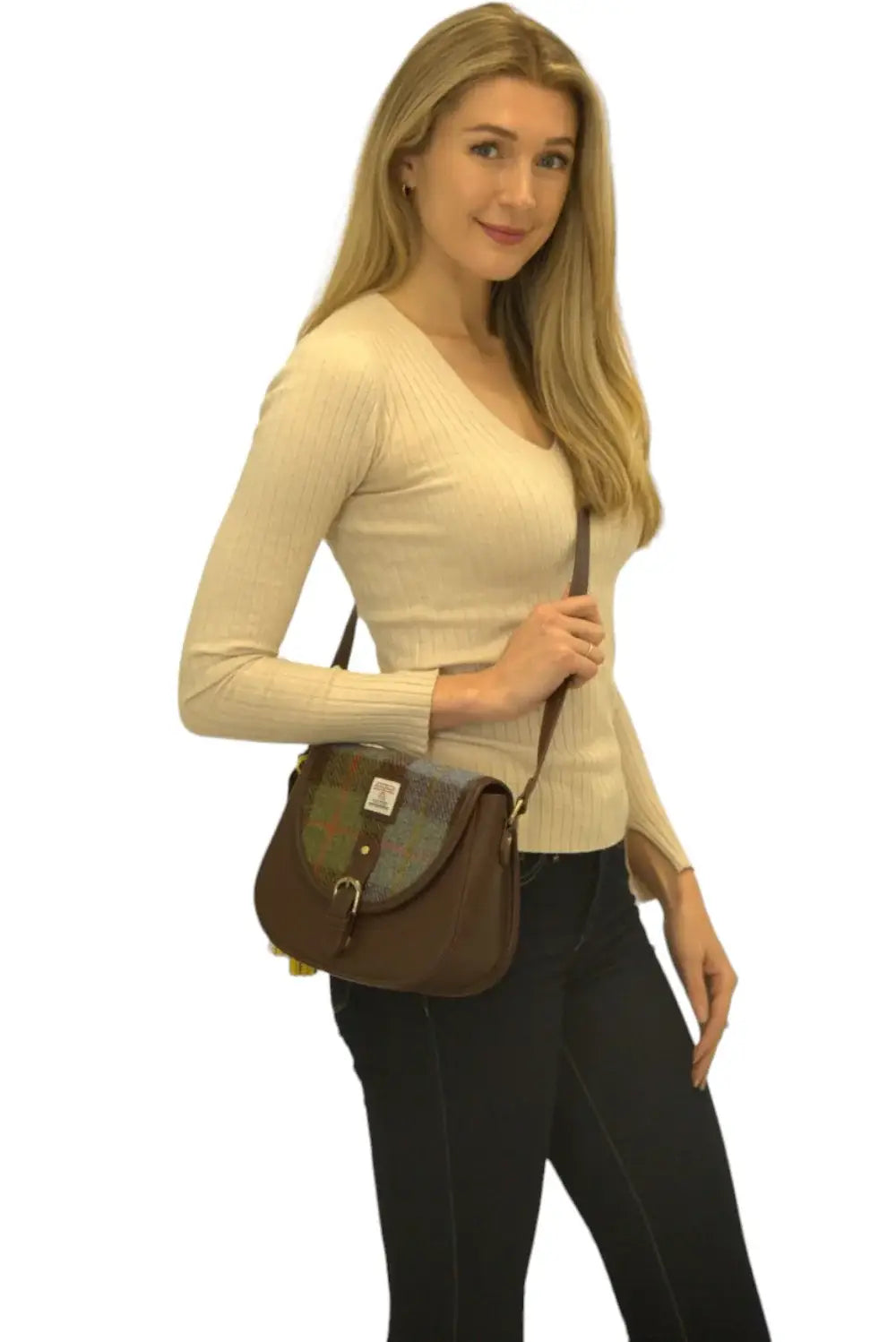 A woman with long blonde hair, wearing a beige sweater and dark jeans, poses against a white background with the Harris Tweed Saddle Bag in dark brown, featuring blue, brown & yellow tartan wool.