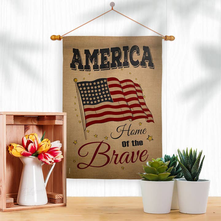 Display the America Home of the Brave Americana Star and Stripes Flag beside a wooden crate with flowers and potted plants for a charming patriotic garden look.