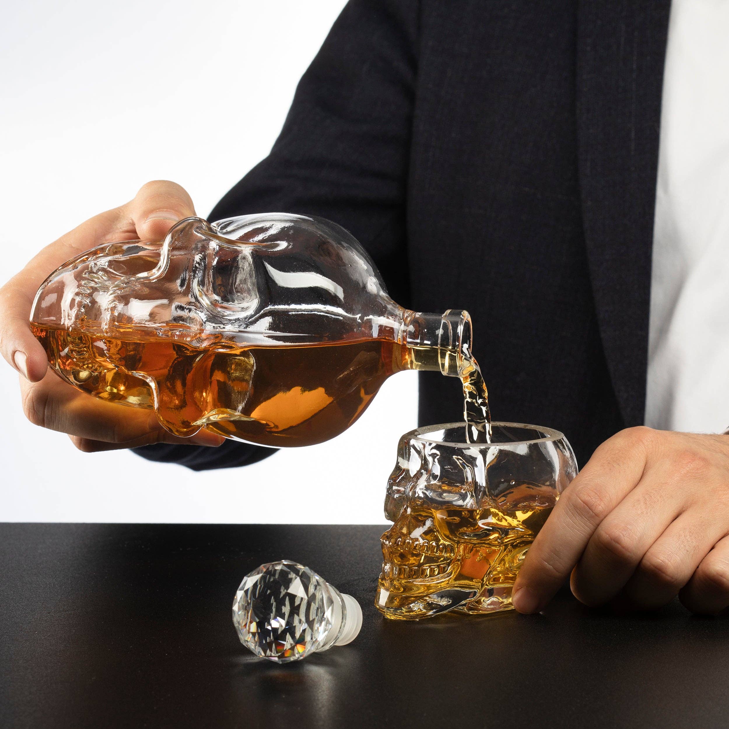 A person pours amber liquid from the Skull Decanter Set into a Skeleton Skull Shot Glass on a black table, perfect for pirate-themed parties. A crystal stopper rests nearby. Includes 1 Skull Decanter & 4 Skeleton Skull Shot Glasses.