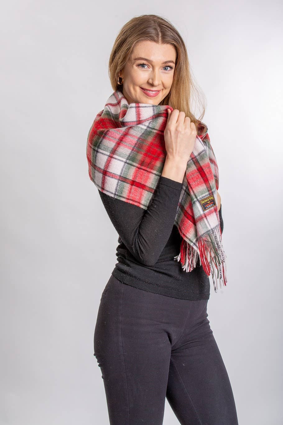 A woman wears the MacDuff Lambswool Scarf in red, white, and green over a black long-sleeve top and black pants, standing against a plain light background.