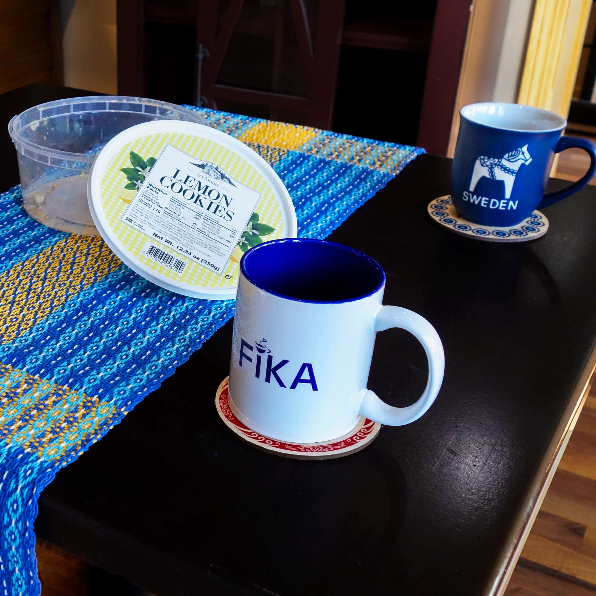 A white FIKA mug and a blue Sweden mug sit on coasters beside an open lemon cookies container on a dark table with a Swedish Flag Runner (65, handwoven on antique loom, 100% cotton) in blue and yellow.