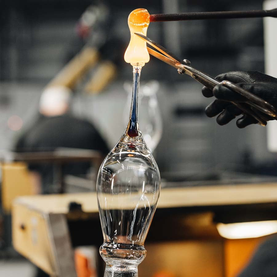 In a glassblowing studio, a person skillfully shapes molten glass with tools, creating an object reminiscent of the elegant Orrefors Intermezzo Blue Flute 7oz.
