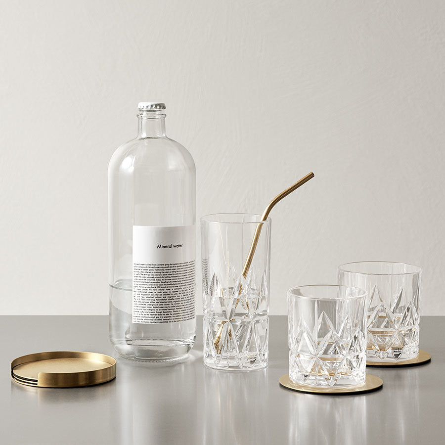 The Orrefors set includes a glass bottle with a metal cap, two empty Peak Double Old Fashioned crystal glasses from the 11.5 oz 4-pack, and one with a metal straw, all elegantly arranged on Martti Rytkönens gold coasters on a sleek gray surface.