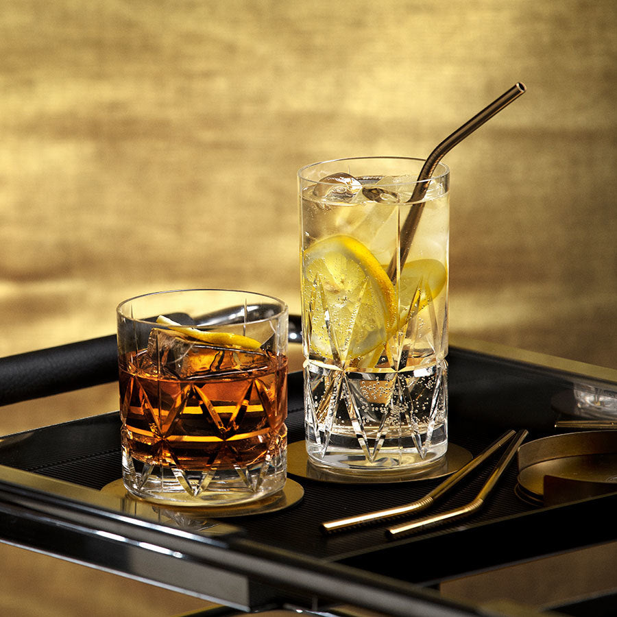 Two drinks on a tray: one in an Orrefors Peak Double Old Fashioned glass with dark liquid and ice, the other in a tall Martti Rytkönen-designed glass with clear liquid, lemon slices, ice, and a metal straw.