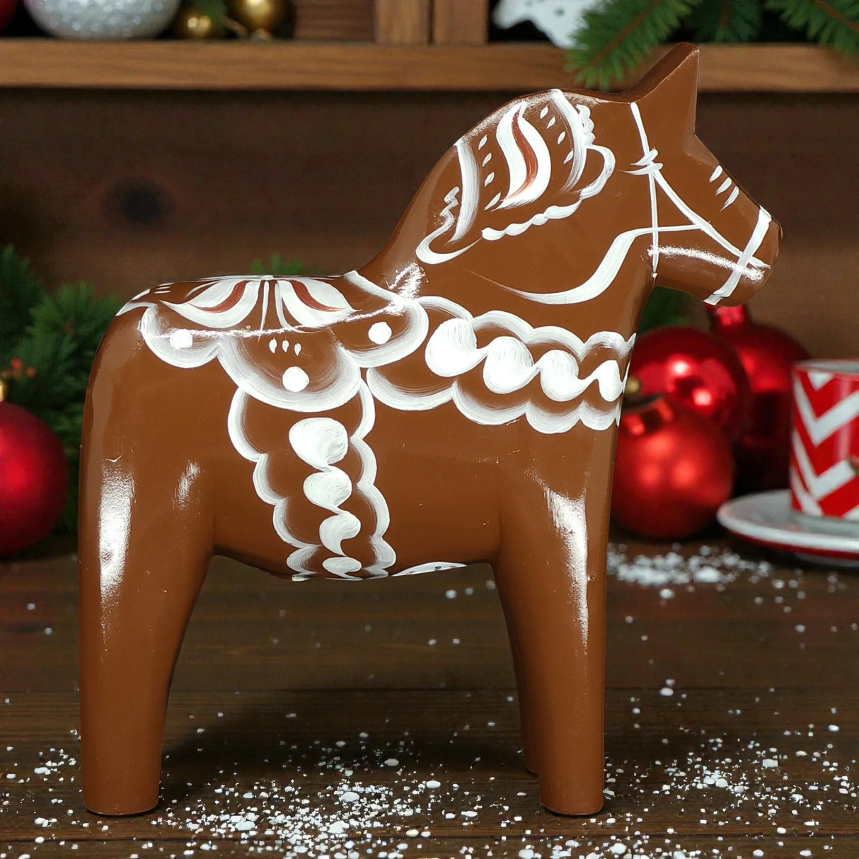 A handcrafted 13 cm Brown Swedish Dala Horse "Gingerbread" by Grannas Olsson features white patterns and stands on wood, accented by red ornaments, pine branches, and a red-and-white cup for a festive Scandinavian touch.
