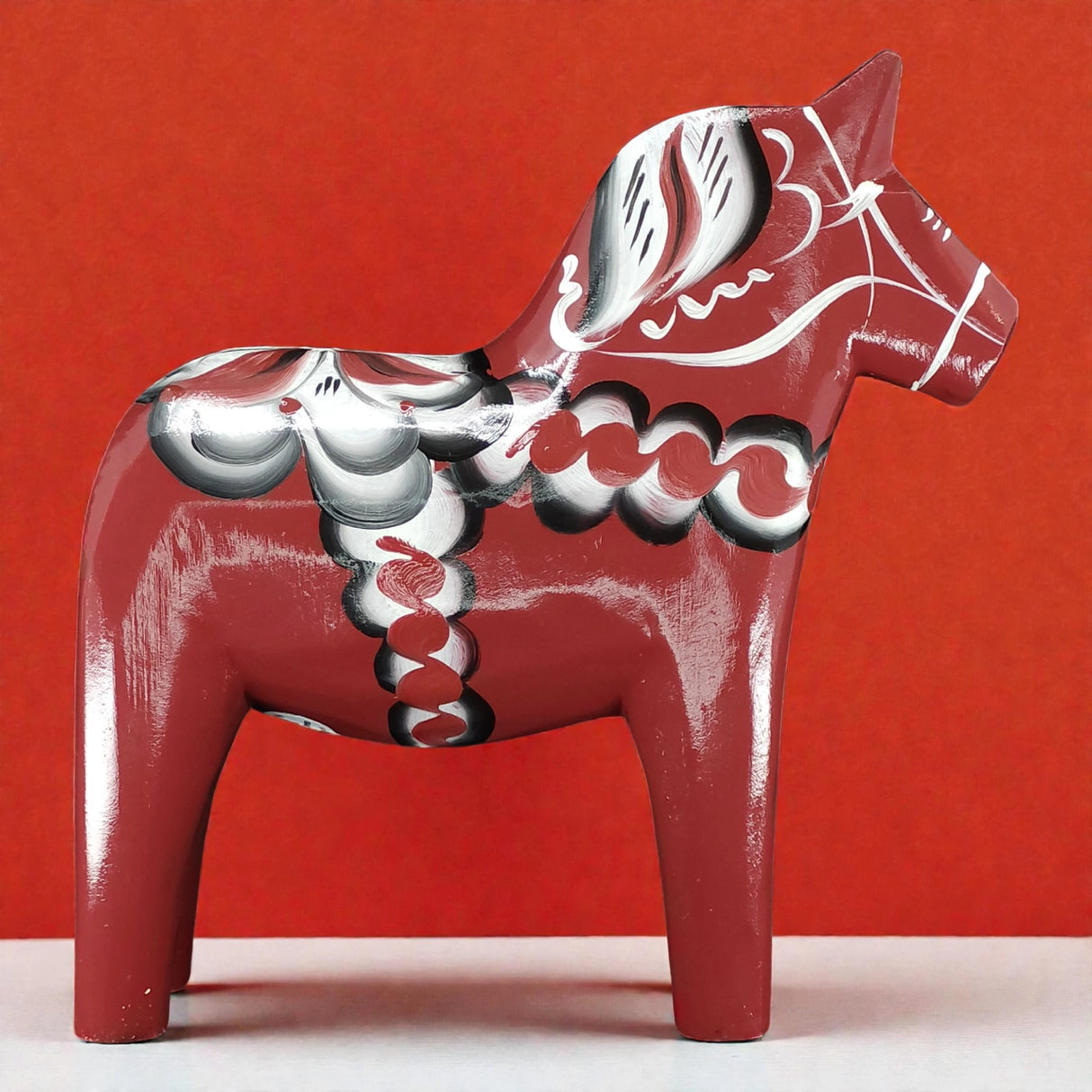 The Grannas Olsson Burgundy Swedish Dala Horse "Maria / Anniversary Series" (15 cm), handcrafted in Sweden, features intricate white and black patterns and stands on a white surface against a red background.