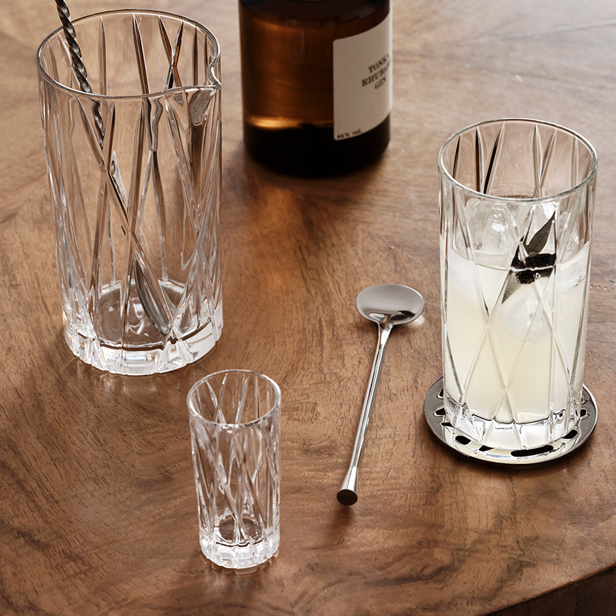 Orrefors: City Highball Glass 12oz (2-Pack) elegantly rests on a wooden table, accompanied by a shaker, mixer, and shot glass. A brown bottle in the background enhances the sophisticated scene.