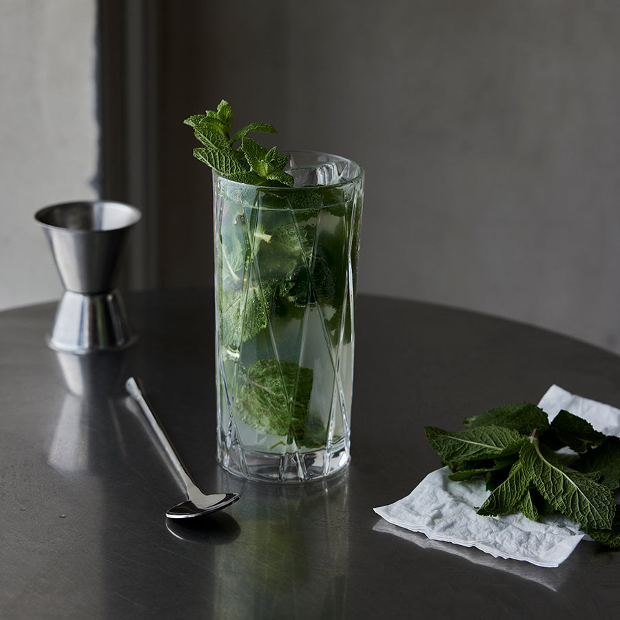 A mojito cocktail in an Orrefors City Highball Glass sits on a metal table, with a spoon, jigger, and mint leaves on a nearby napkin.