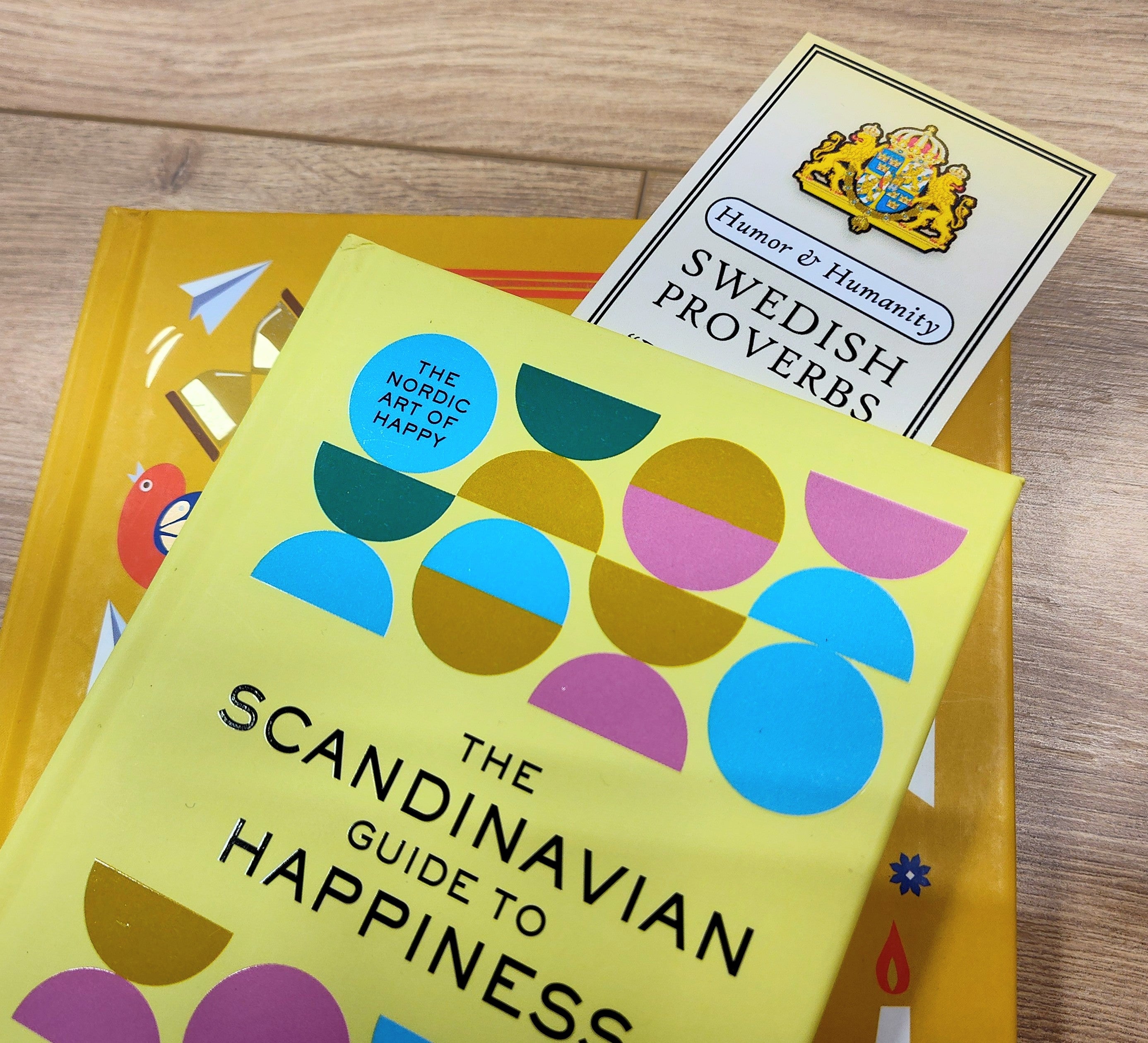 The Scandinavian Guide to Happiness, paired with a Bookmark: Swedish Proverbs “Humor & Humanity” (2.5" × 8.5"), sits on a yellow book atop wood—showcasing the warmth and wit of Scandinavian tradition.