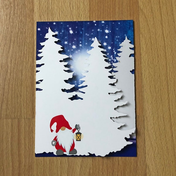 The Snowy Pines Nisse Winter Card features a laser-cut design of a Norwegian Nisse with a lantern, surrounded by snowy pines and set against a starry night sky.