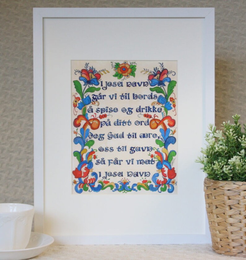 Framed artwork titled I Jesu Navn by Greta Lann, featuring Norwegian Table Grace with floral and scroll designs, placed on a tabletop beside a white cup and a basket with a plant.