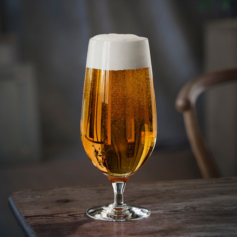 A 20oz Orrefors Beer Lager glass from the Erika Lagerbielke-designed 4-pack holds golden beer with a foamy head, resting on a wooden table in a softly lit room.