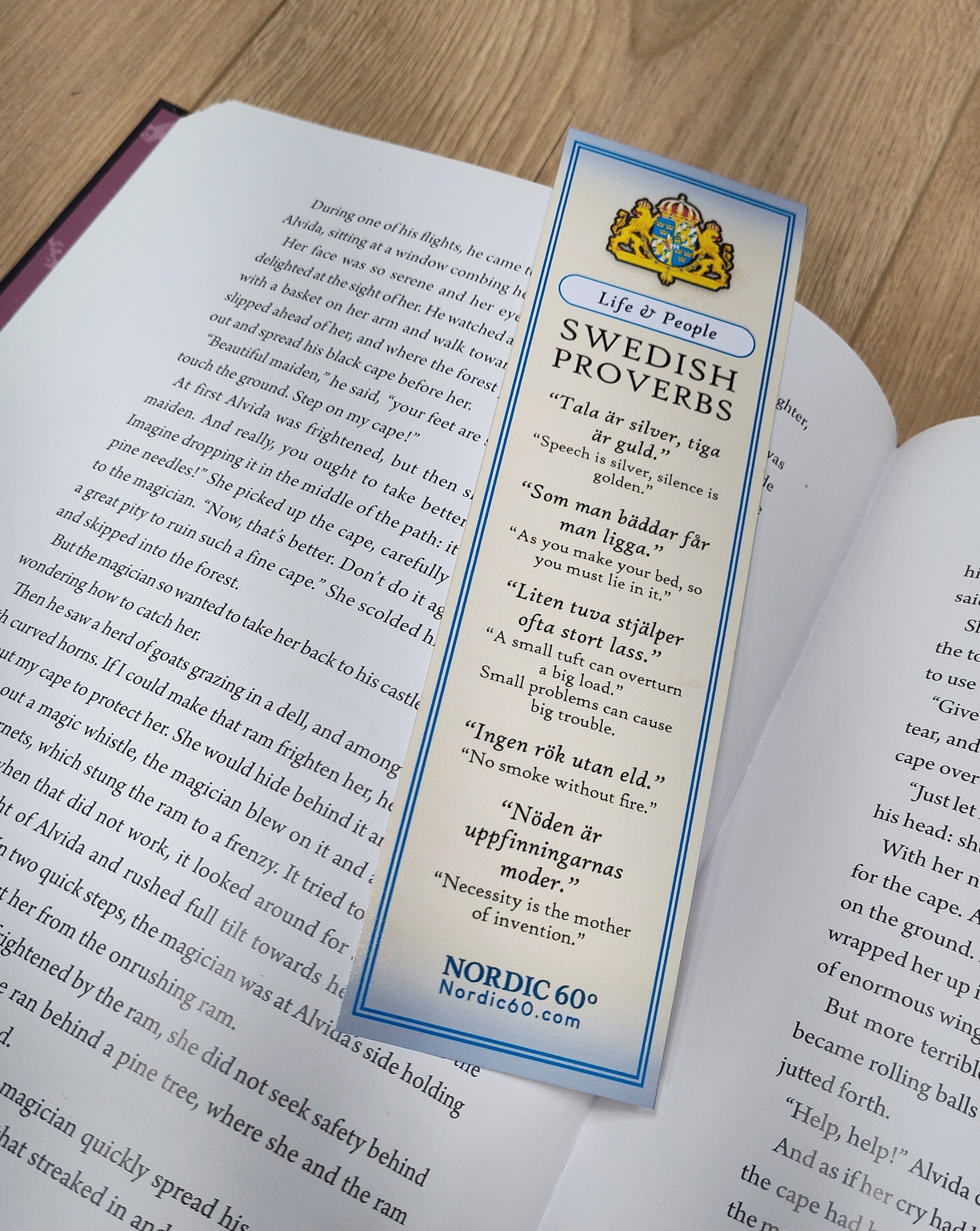 The Bookmark: Swedish Proverbs “Life & People” (2.5" × 8.5") with blue and white Scandinavian design rests on the open pages of an English book placed on a wooden surface.