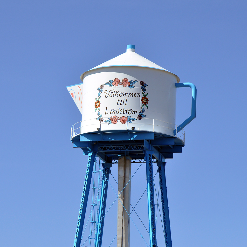 A whimsical Nordic sticker of Lindstroms Swedish Coffee Pot Water Tower, featuring floral designs and Välkommen till Lindström, captures the charm of Minnesotas clear blue sky.