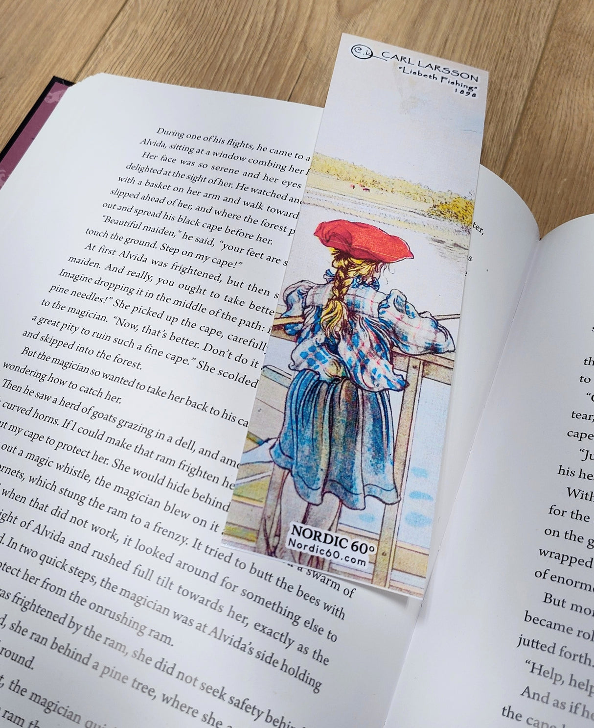 A book rests open on wood, marked by the Carl Larsson “Lisbeth Fishing” bookmark (8.5" × 2.5"), which showcases Lisbeth in her red hat and captures the essence of a Swedish summer.