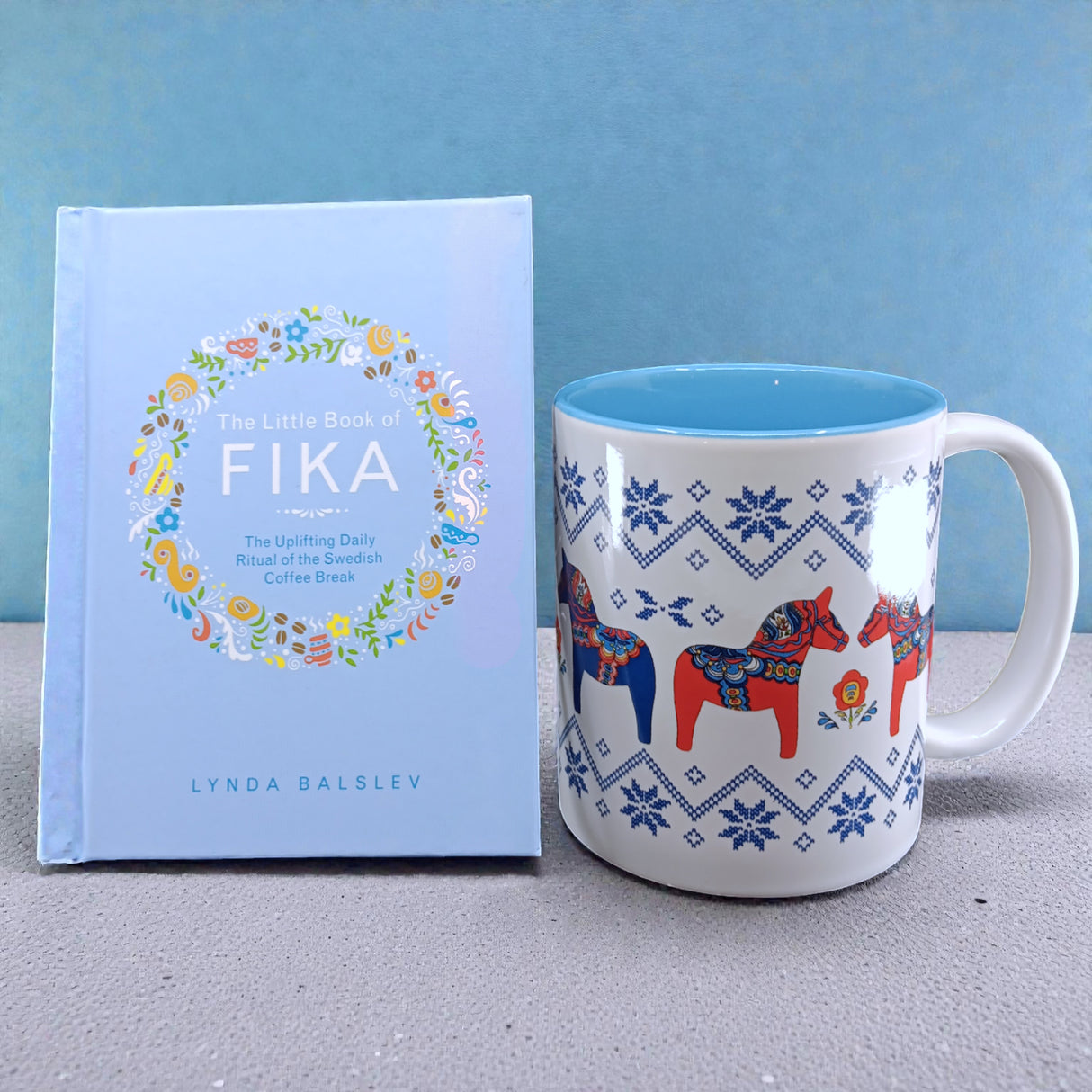 A light blue copy of "Little Book of Fika" by Lynda Balslev stands beside a white mug with blue patterns and red Dala horses, capturing the essence of a Swedish coffee break, all set against a blue background.