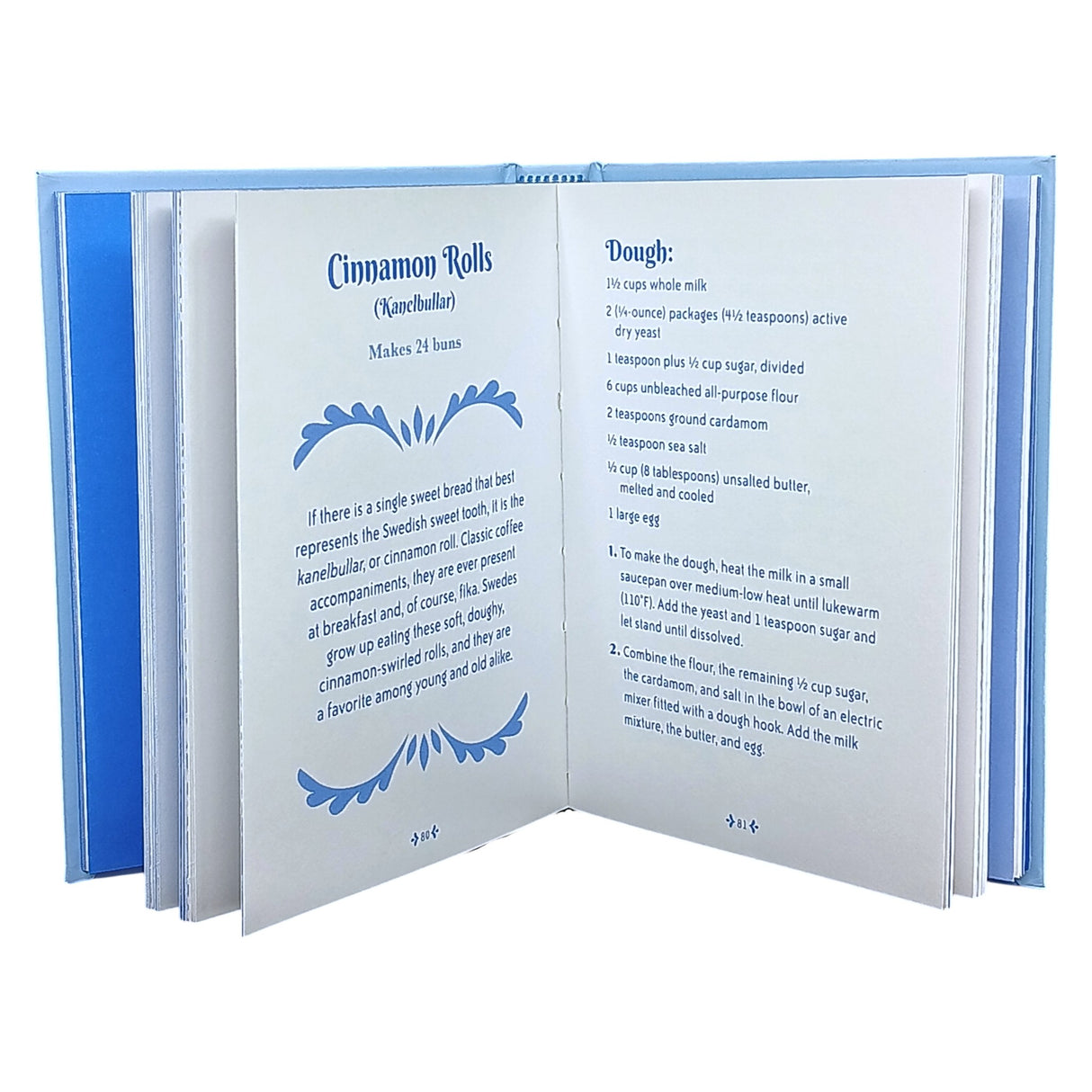 The open Little Book of Fika by Lynda Balslev shows a cinnamon rolls recipe with ingredients and instructions across two pages, ideal for a Swedish coffee break, and features a blue and white decorative border on the left page.