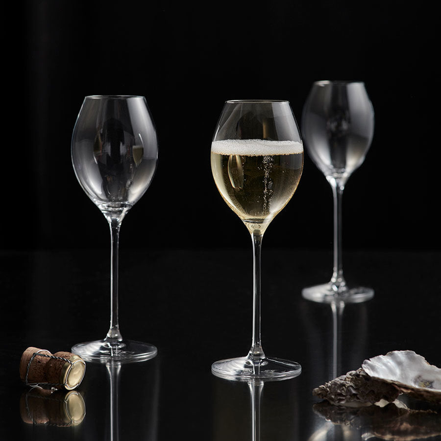 Three Orrefors wine glasses sit on a reflective surface; one filled with champagne, two are empty. Nearby, a More Champagne Boule Glass by Erika Lagerbielke shines as a cork and oyster shells lie in elegant disarray.