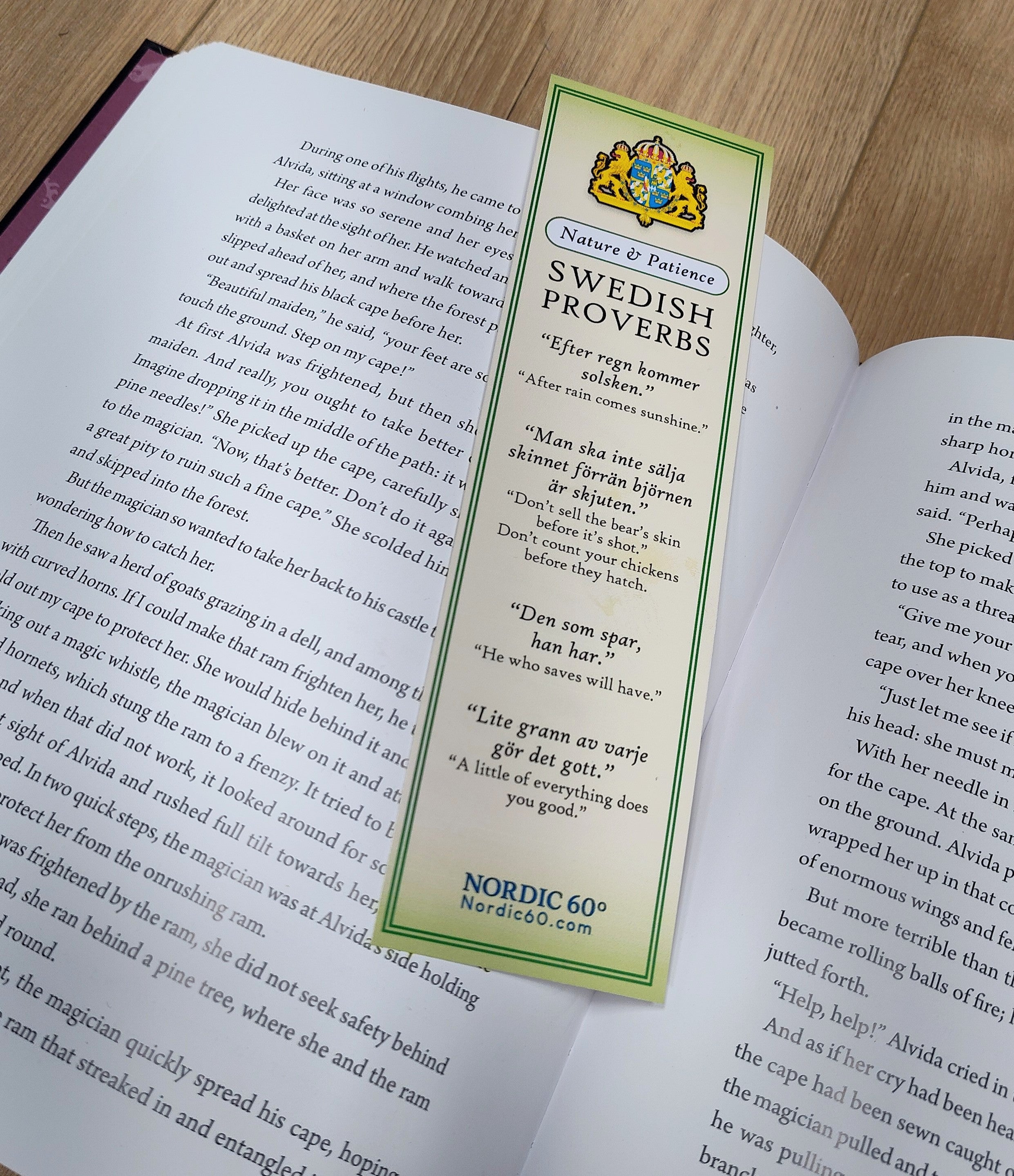 The Bookmark: Swedish Proverbs “Nature & Patience” (2.5" × 8.5") lies on the open pages of a book atop a wooden surface, highlighting its green nature-inspired design and wise Swedish sayings.